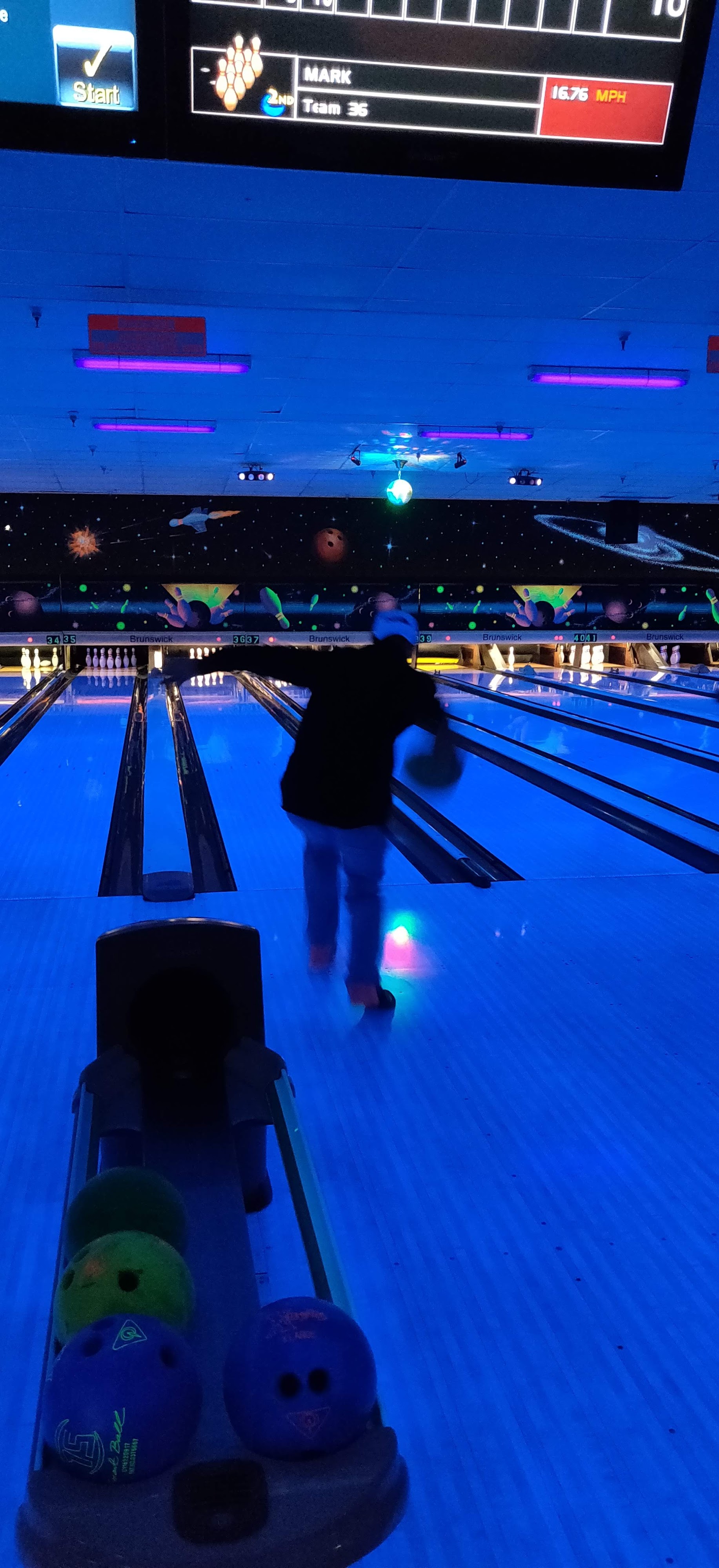 A bowler prepares to release a ball on a blue-lit cosmic bowling lane with space-themed murals.