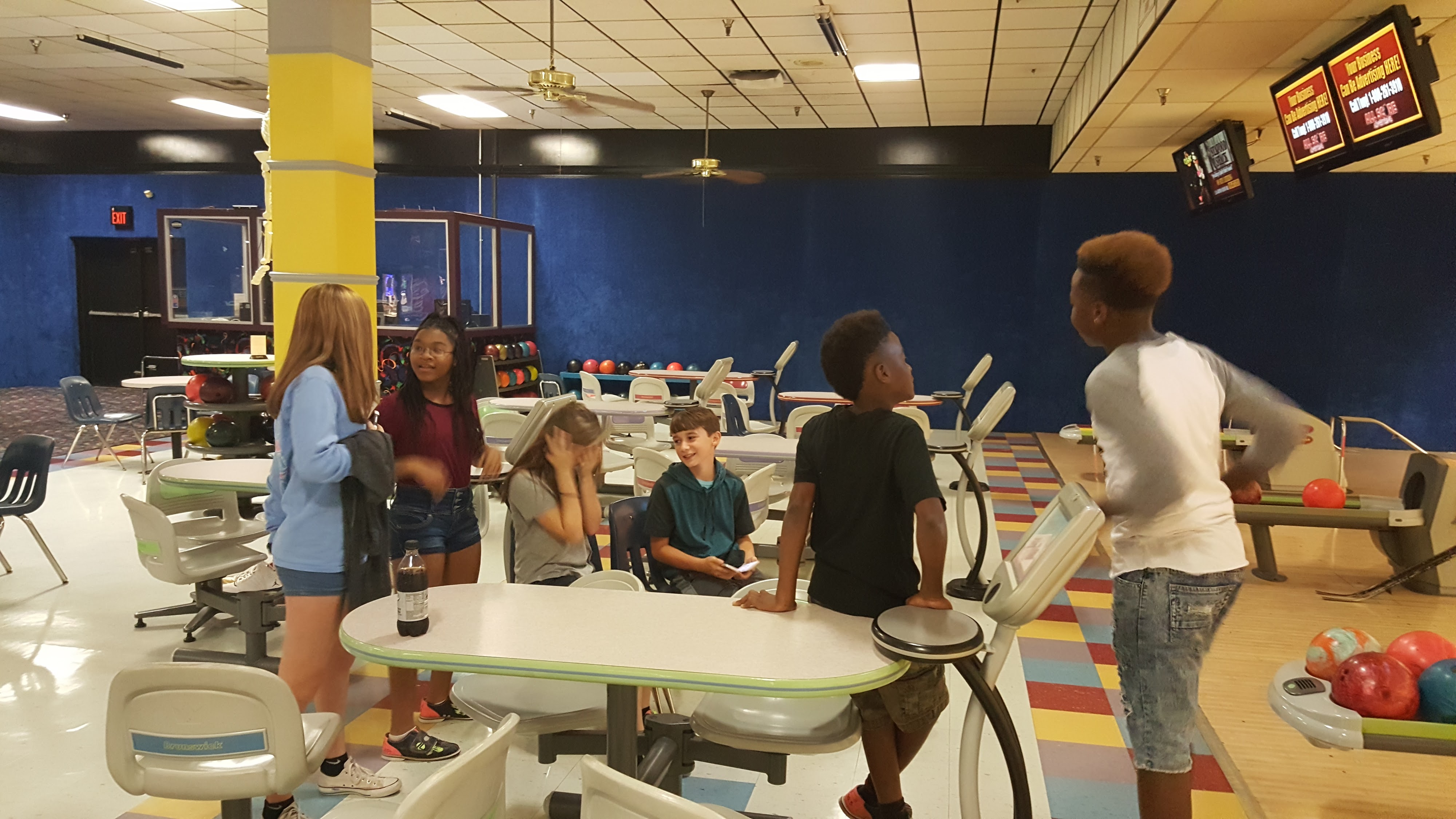 A group of kids and teenagers talk and laugh at tables near the bowling lanes in a brightly lit center.