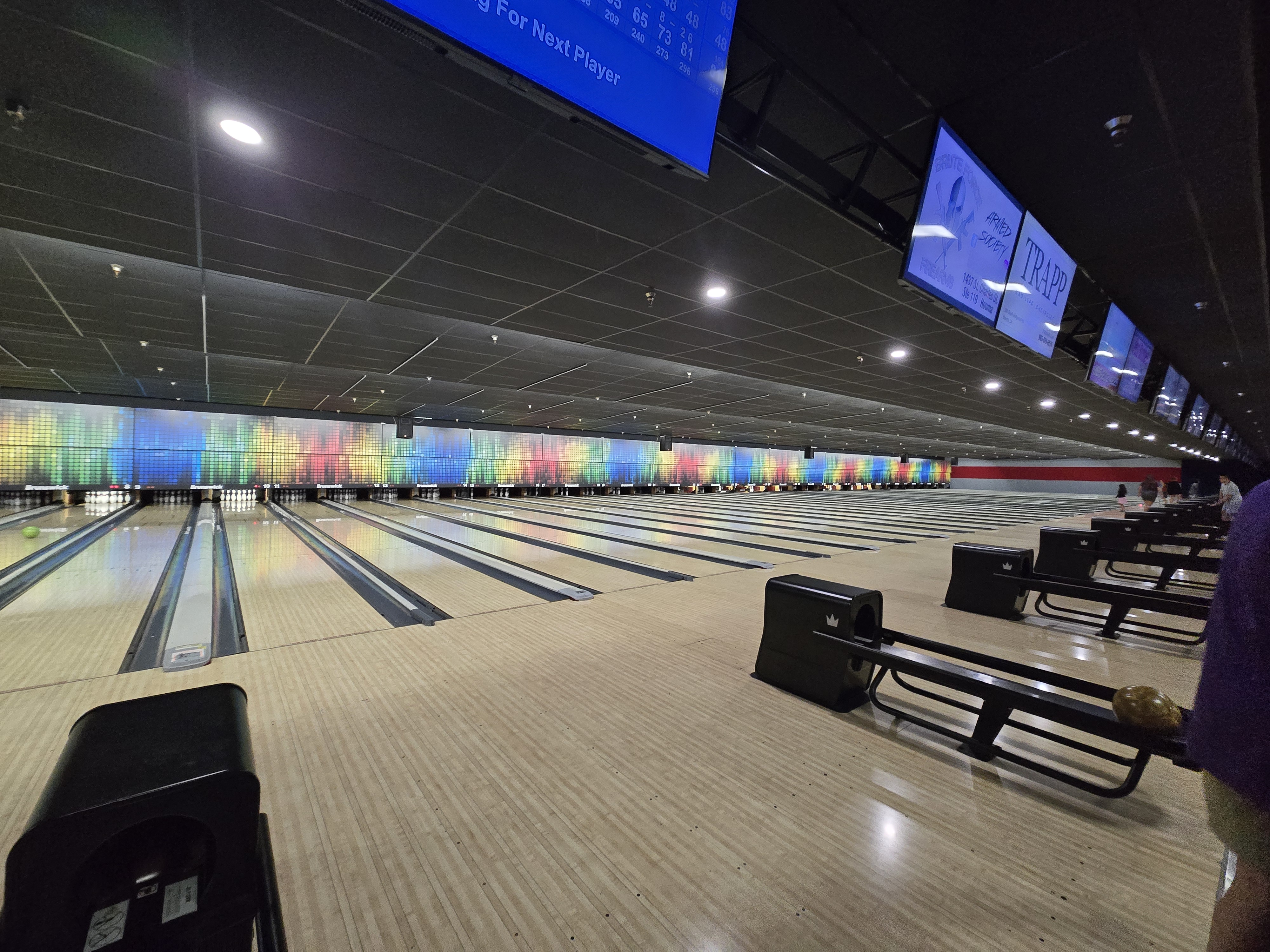 A wide view of several bowling lanes with colorful rainbow backdrops and overhead scoring monitors at Creole Lanes.