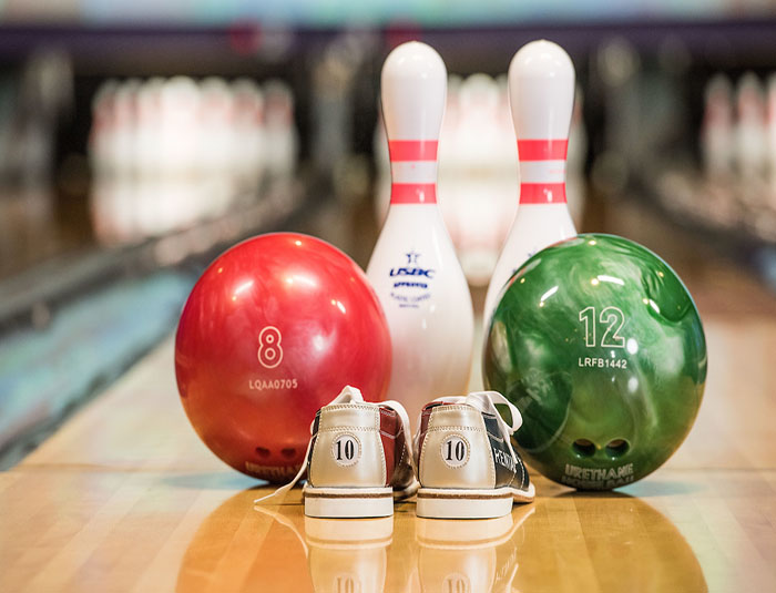 A red and green bowling ball sit next to pins and rental shoes on a wooden lane.