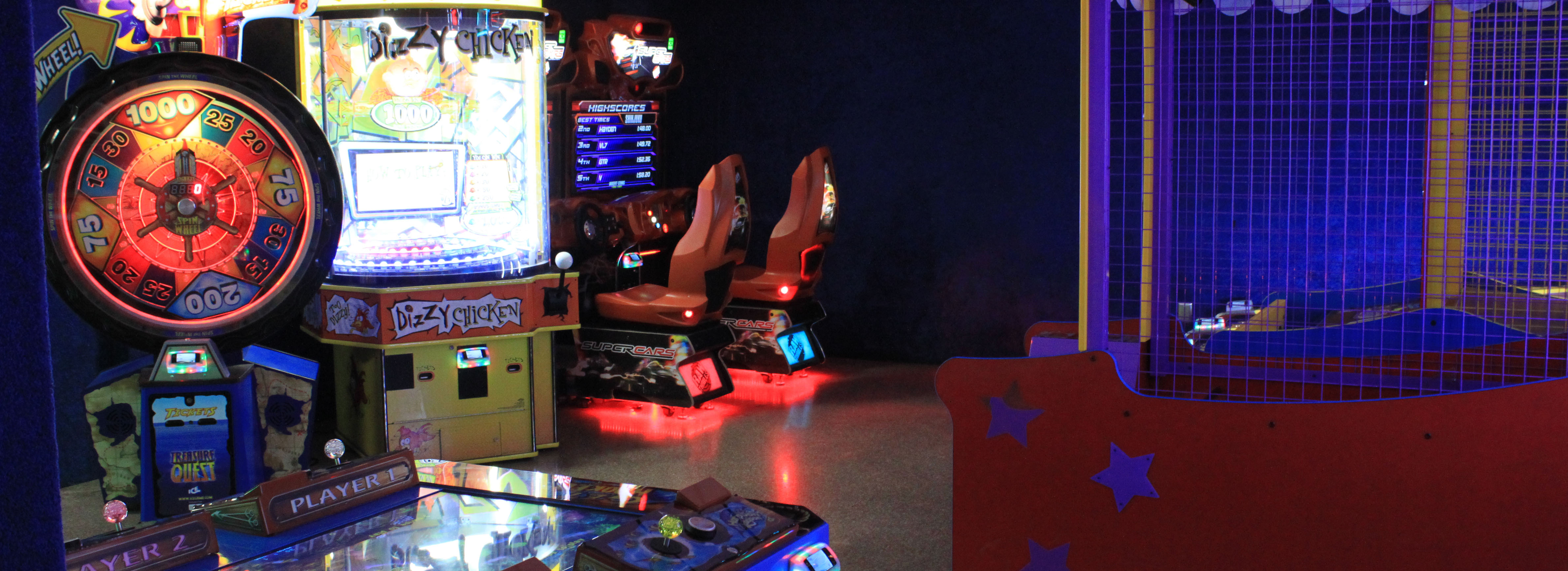 A wide view of a dimly lit arcade featuring a large prize wheel, racing simulators, and ticket redemption games.