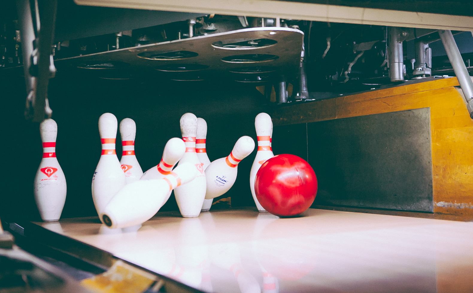 A red bowling ball impacts white pins on a lane, capturing a strike in motion with pinsetter machinery above.