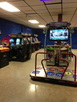 An arcade room with a dance game platform in the foreground and a row of gaming cabinets along a blue wall.