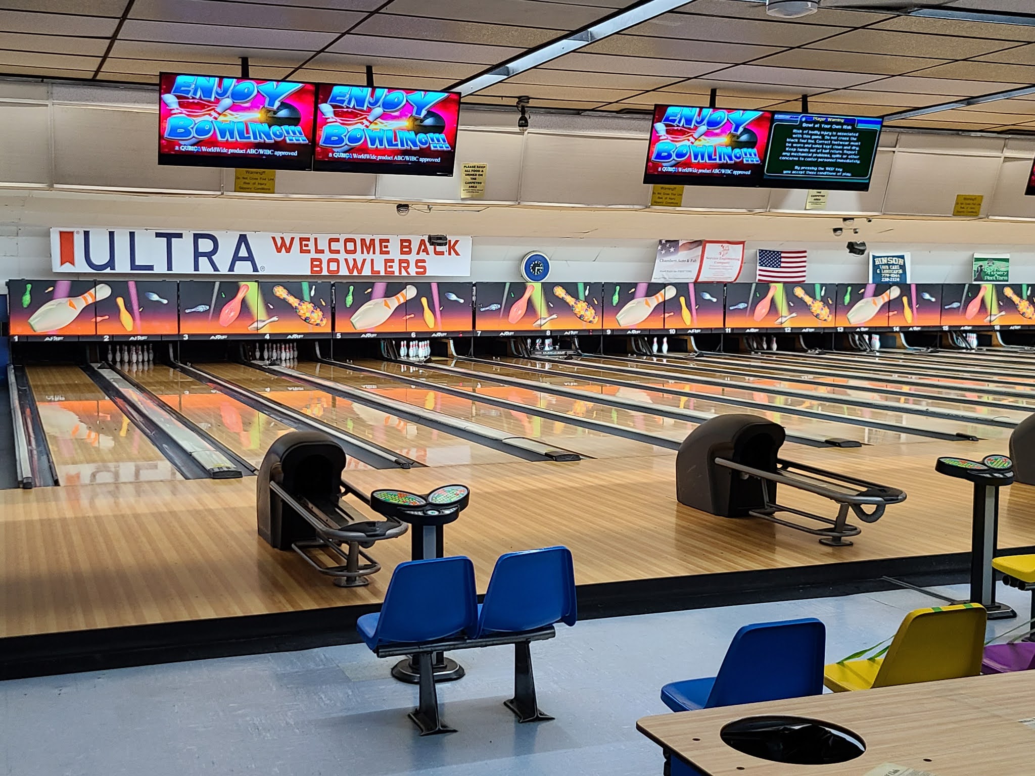 A row of polished wooden bowling lanes with blue and yellow chairs and overhead scoring monitors.