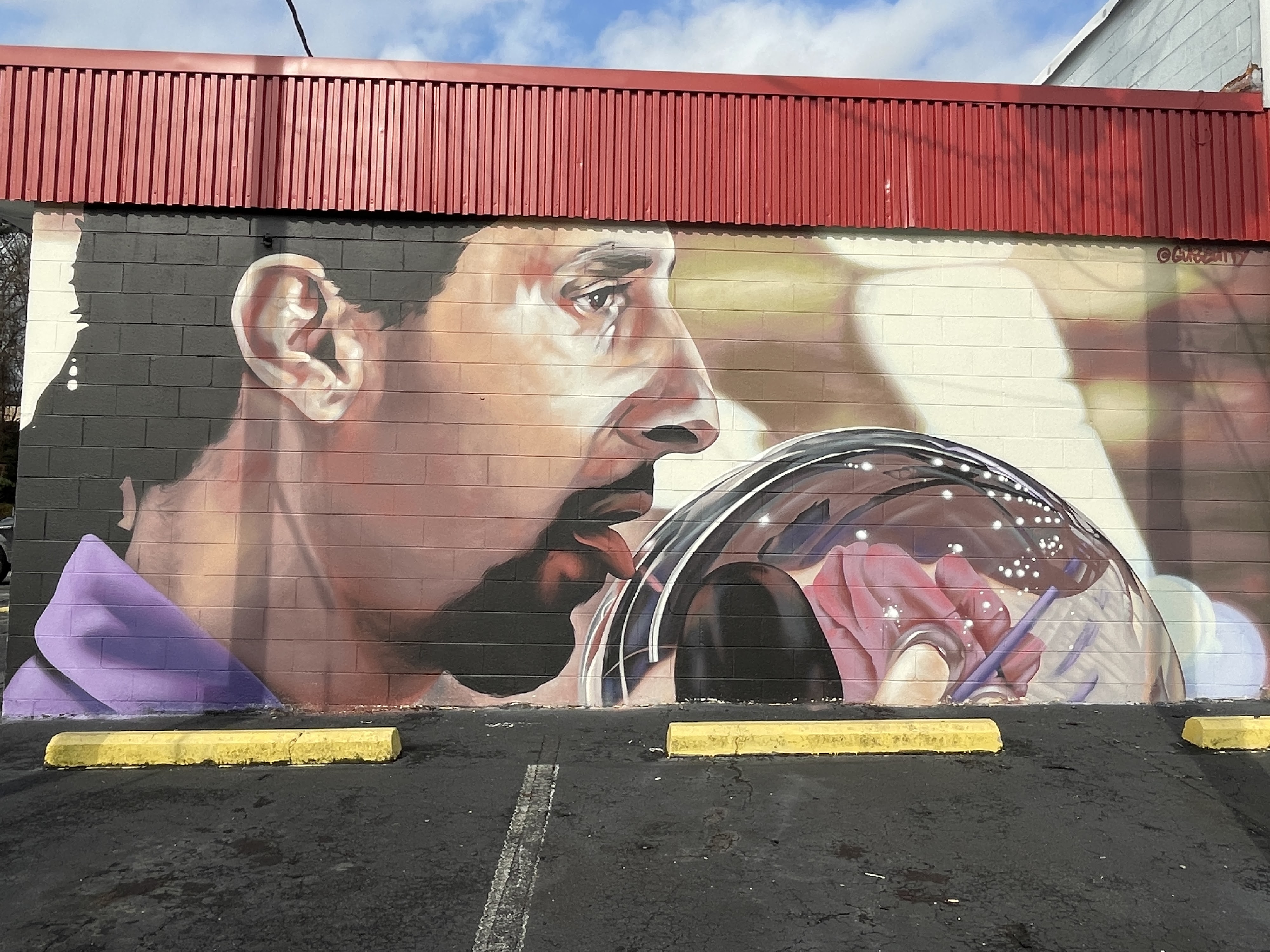 A large outdoor mural of a man in a purple shirt licking a bowling ball on a brick wall.