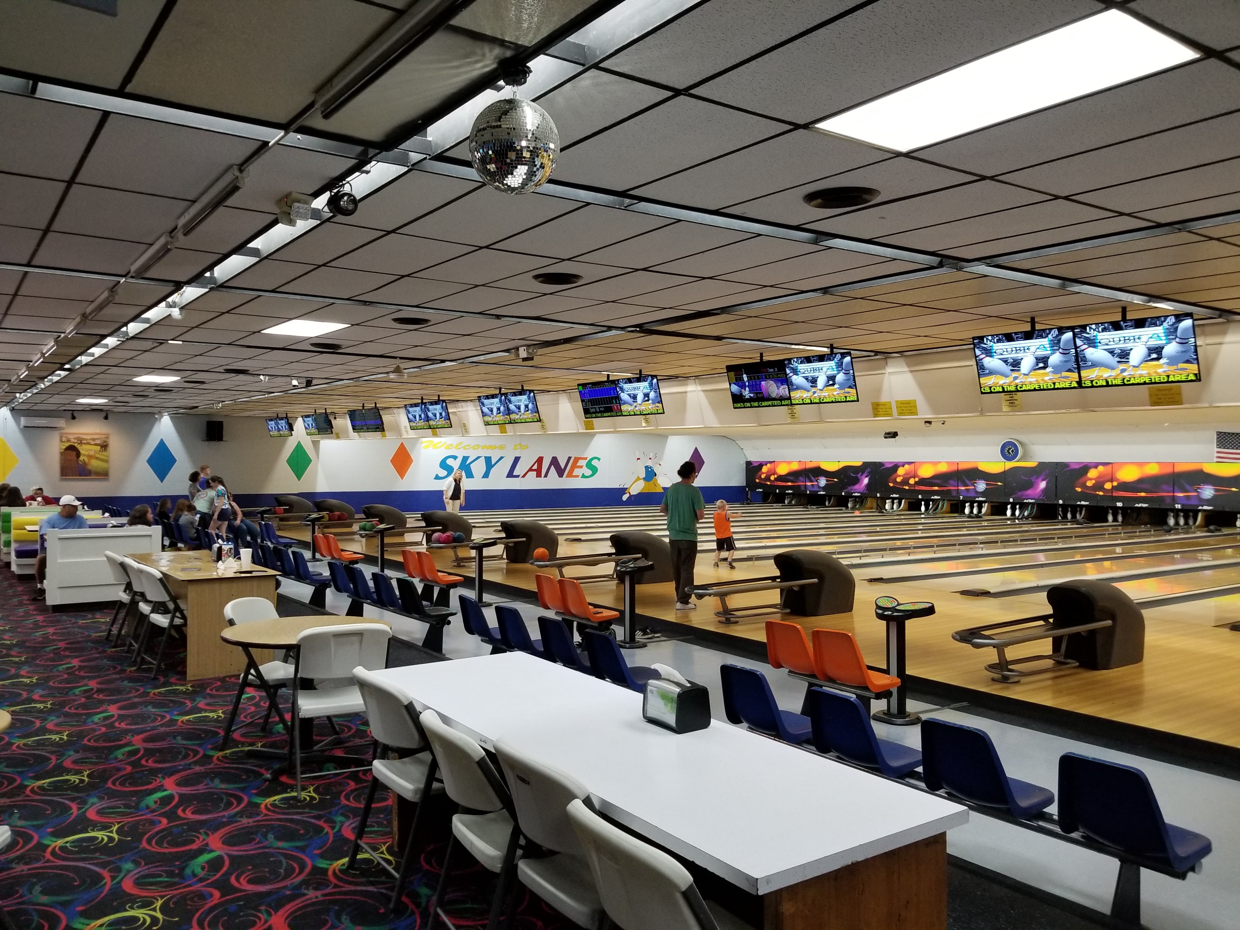 Wide interior view of Sky Lanes Bowling showing multiple lanes, colorful neon swirl carpet, and a disco ball.