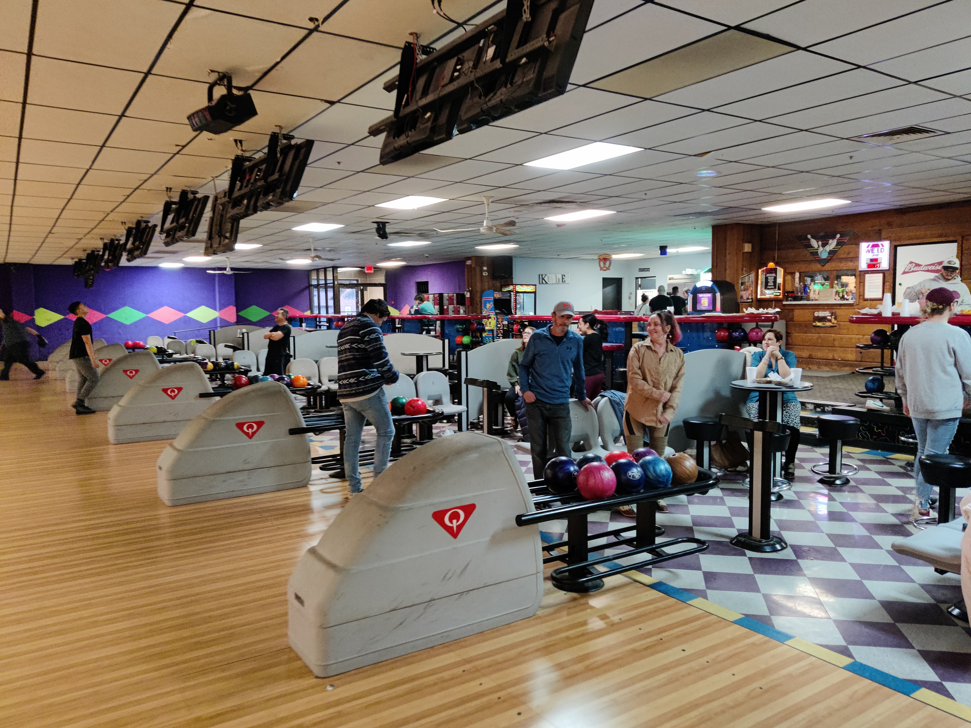 People socialising and preparing to bowl at a bright, modern bowling center with wood lanes and checkered flooring.