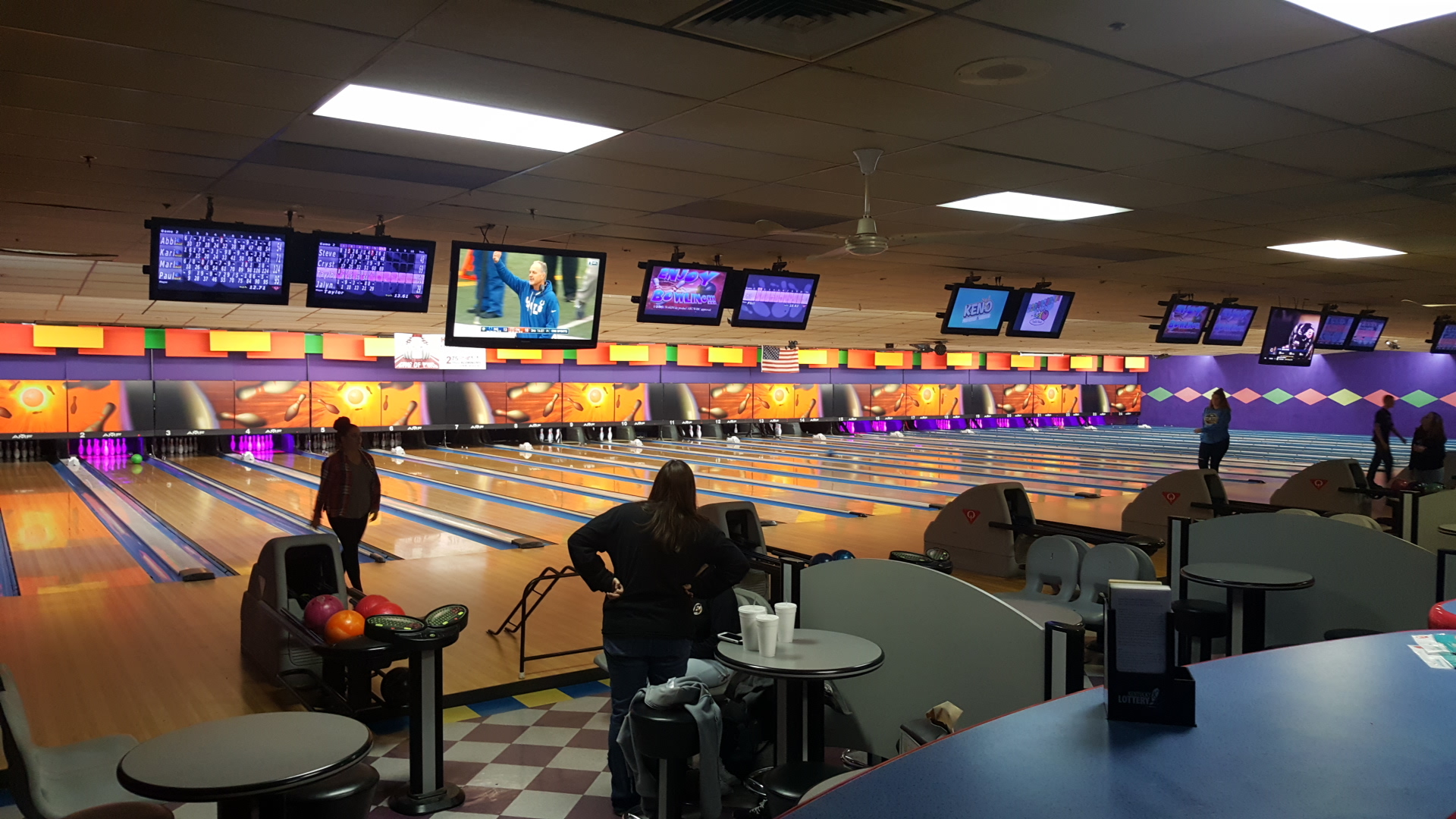 People bowling at a large indoor bowling center with purple accents and overhead scoring monitors.