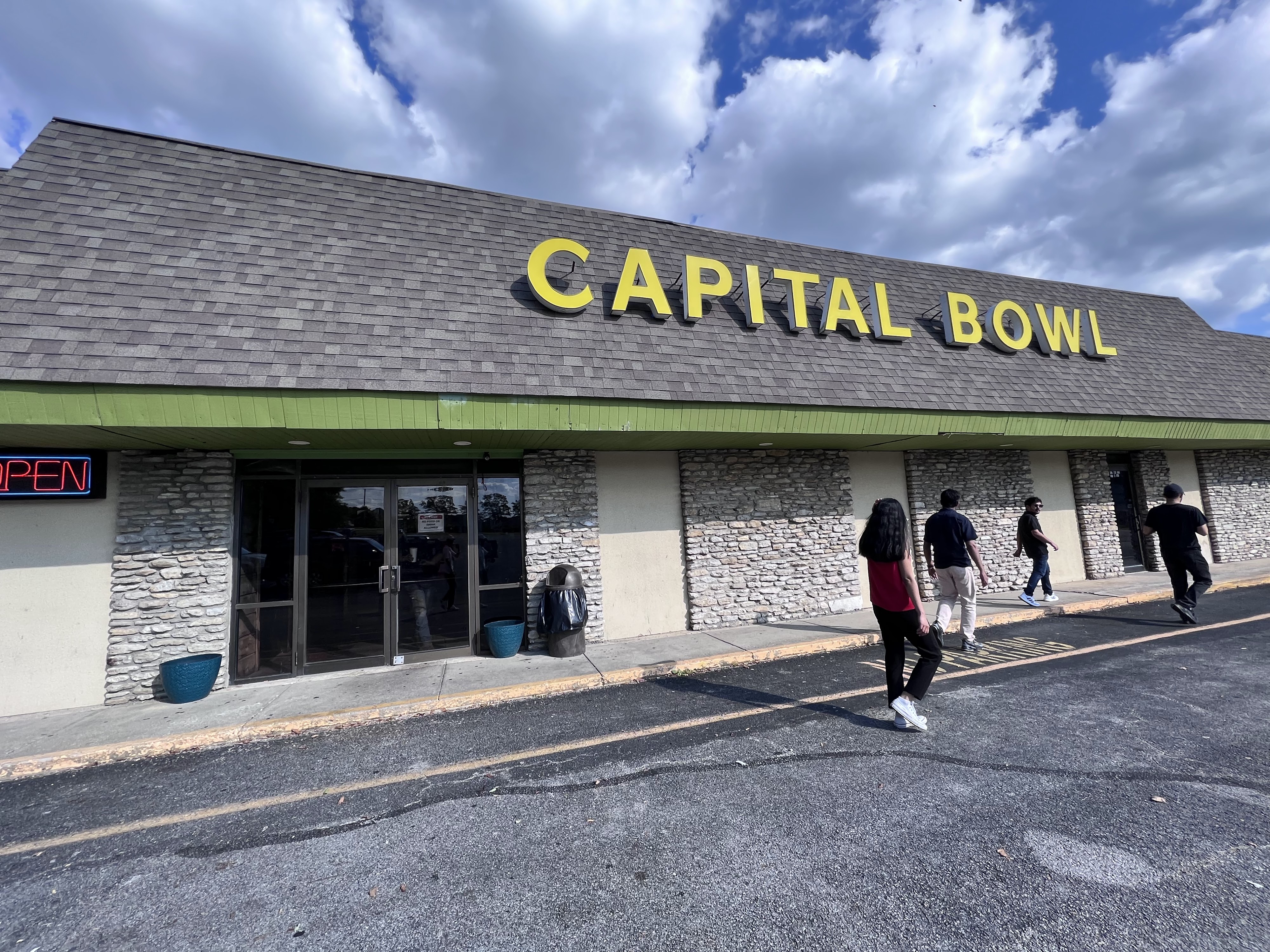 Exterior of Capital Bowl featuring a stone facade and yellow signage under a blue sky with people walking by.