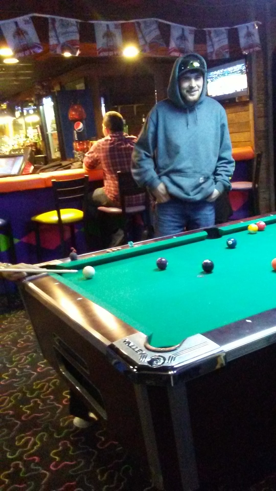A man smiles behind a billiards table in a dimly lit bar area with people sitting on stools in the background.