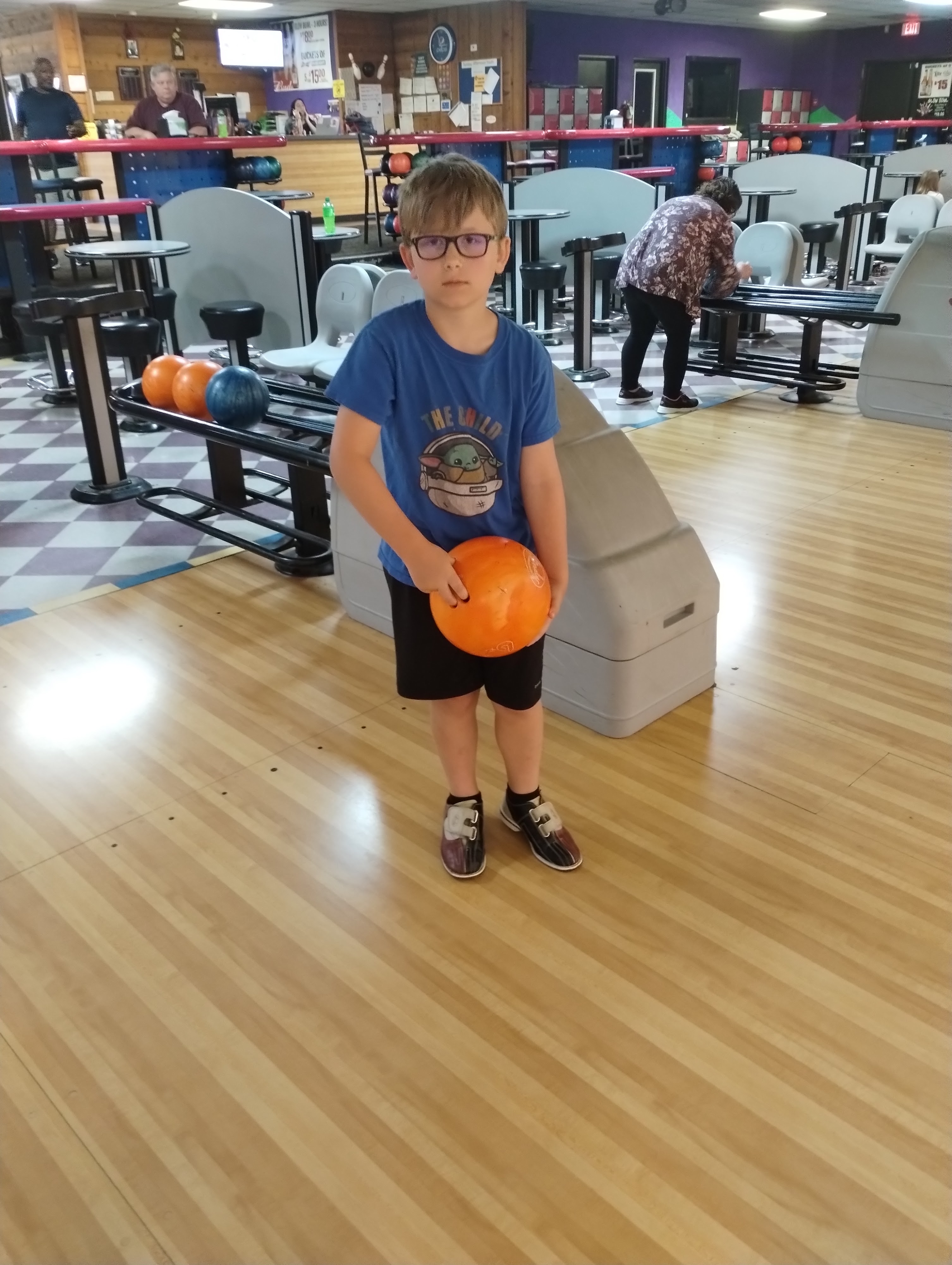 A young boy in a blue shirt and glasses holds an orange bowling ball on a lane at Capital Bowl.