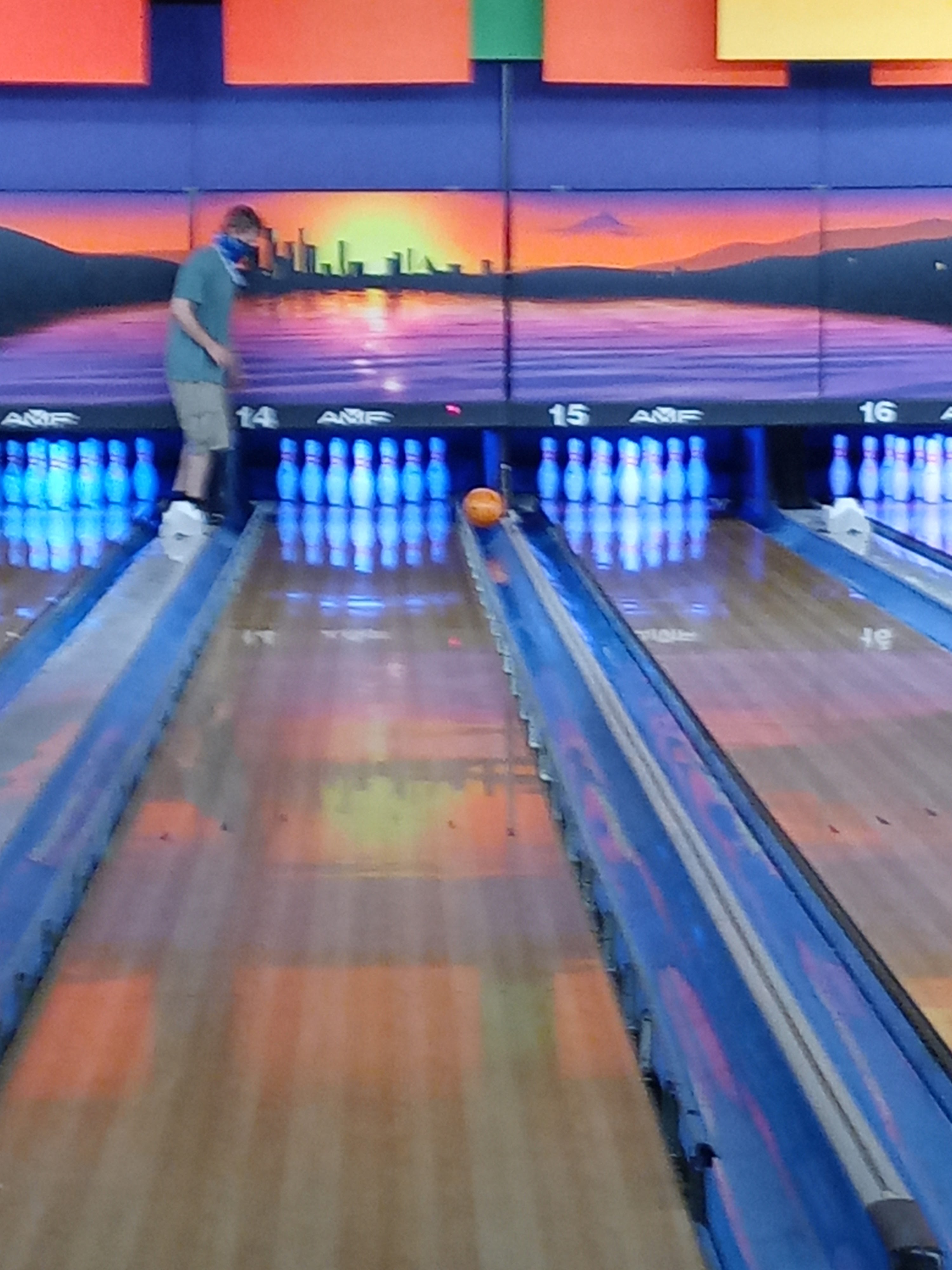 A bowling ball rolls down a lane with bumpers toward white pins under blue and orange lighting.