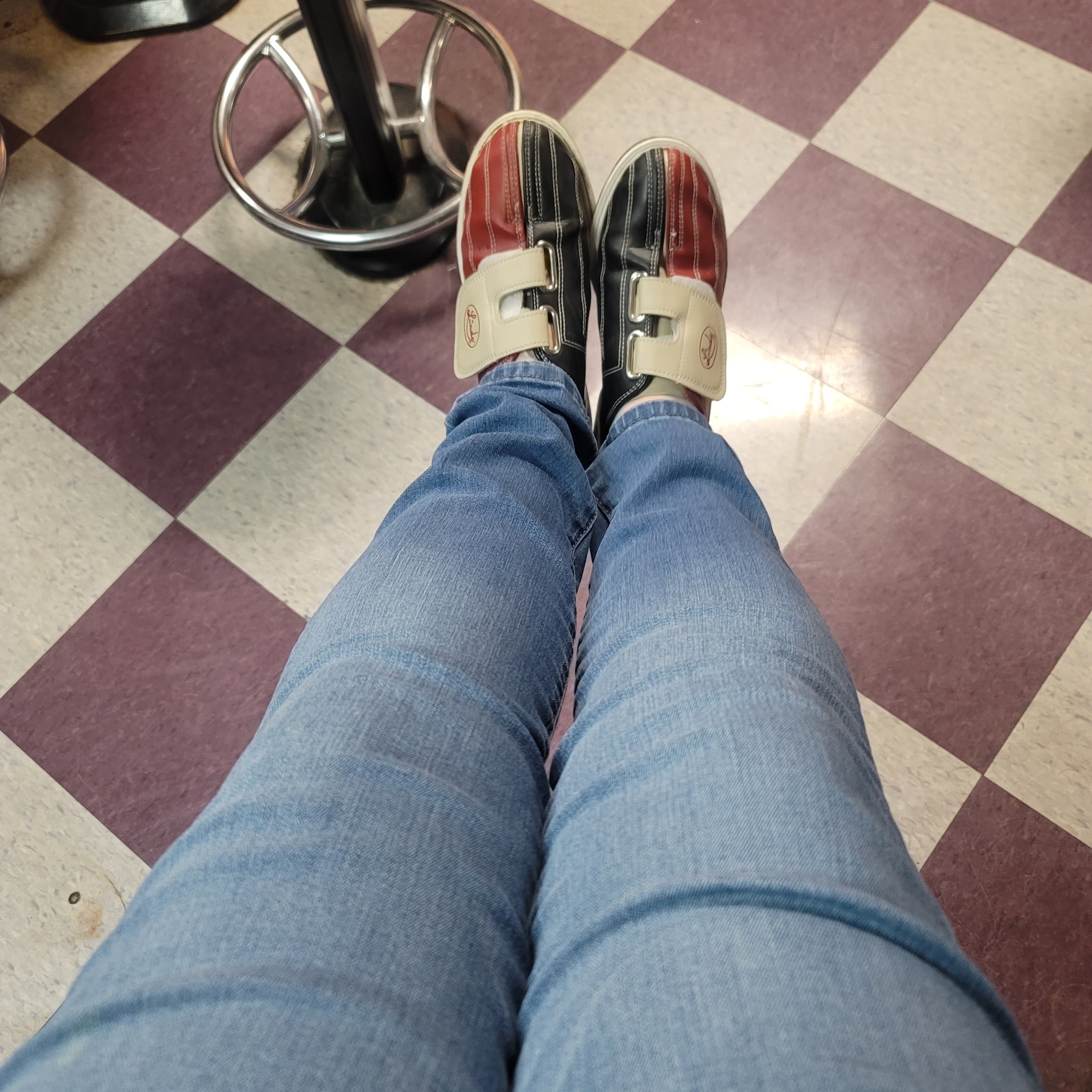 A person wearing red and black rental bowling shoes sitting on a checkered floor.