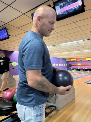 A man in a blue shirt holds a bowling ball while standing on the lane approach at a bowling alley.
