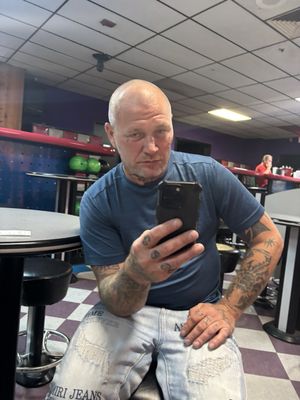 A man with arm tattoos takes a selfie while sitting at a table in a bowling alley.