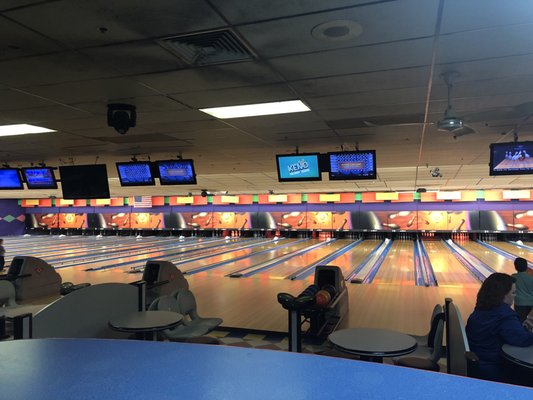 An indoor view of a bowling alley with many lanes, overhead scoring monitors, and casual seating areas.