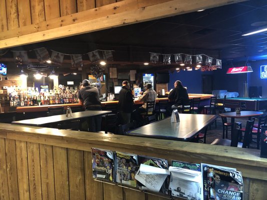 A rustic bar area with patrons seated at the counter and a billiards table in the background.