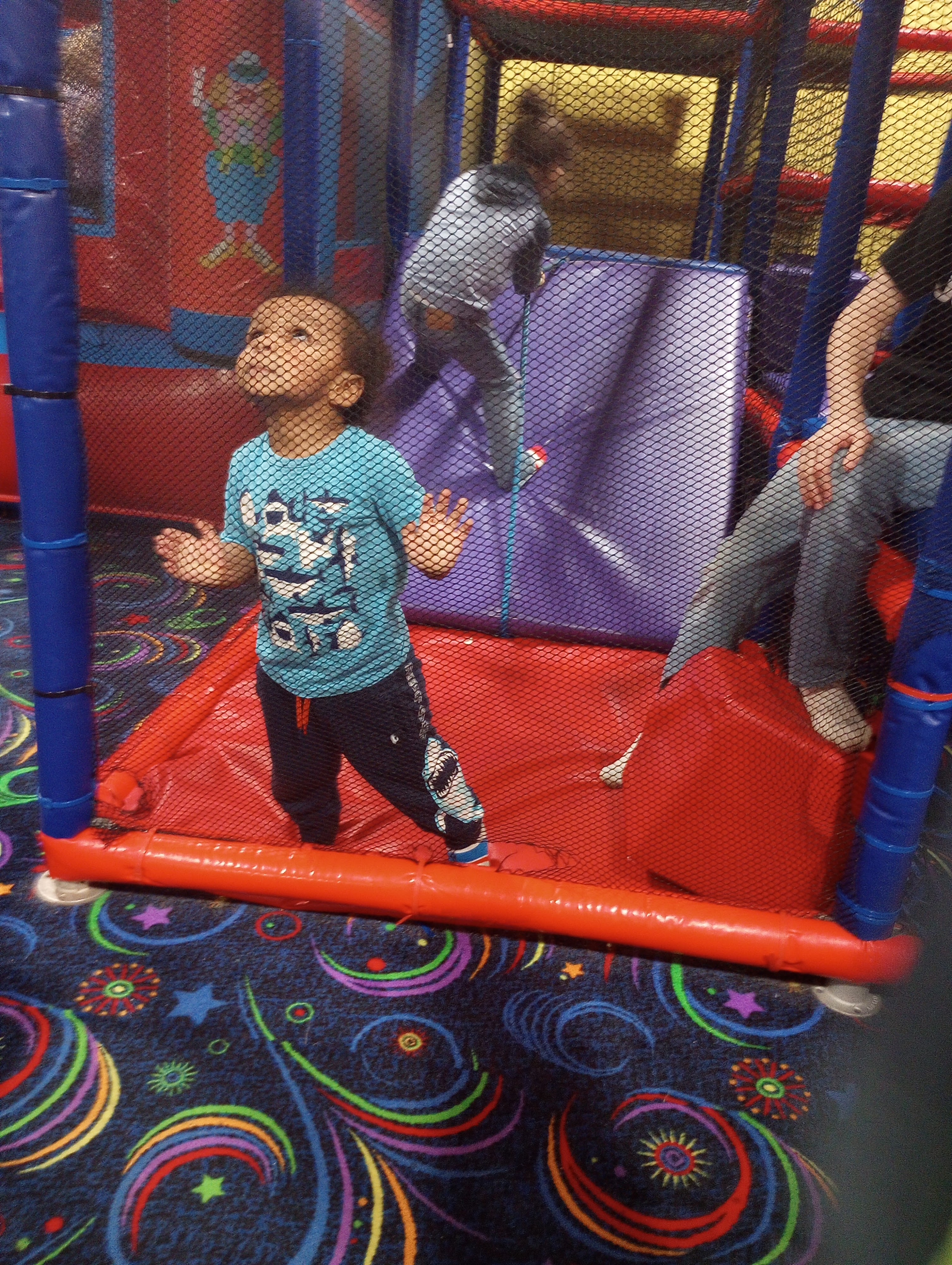 A young child stands inside a multi-level soft play structure with safety netting and colorful padded ramps.