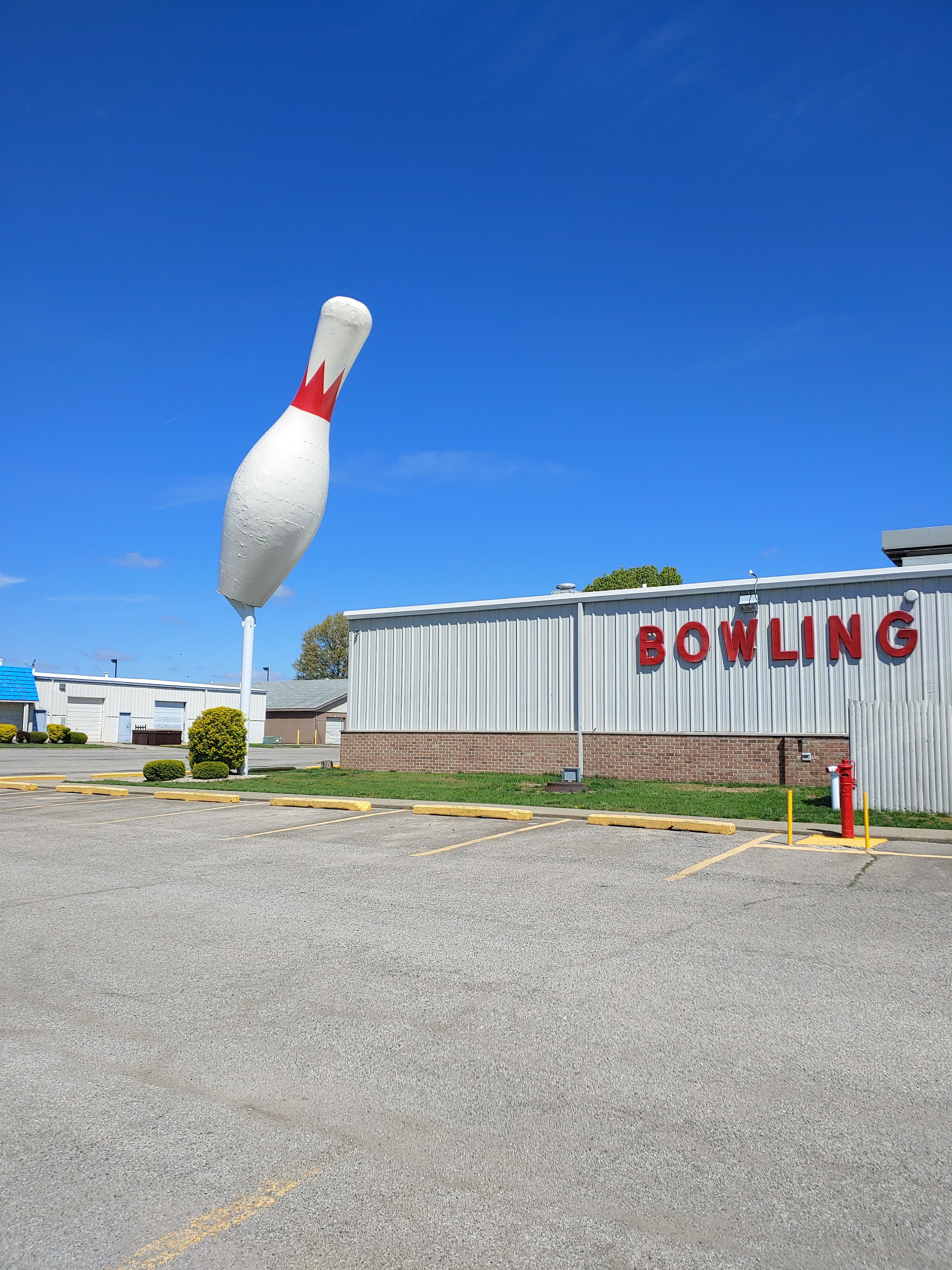A giant white bowling pin statue stands in the parking lot in front of a grey building with red BOWLING text.