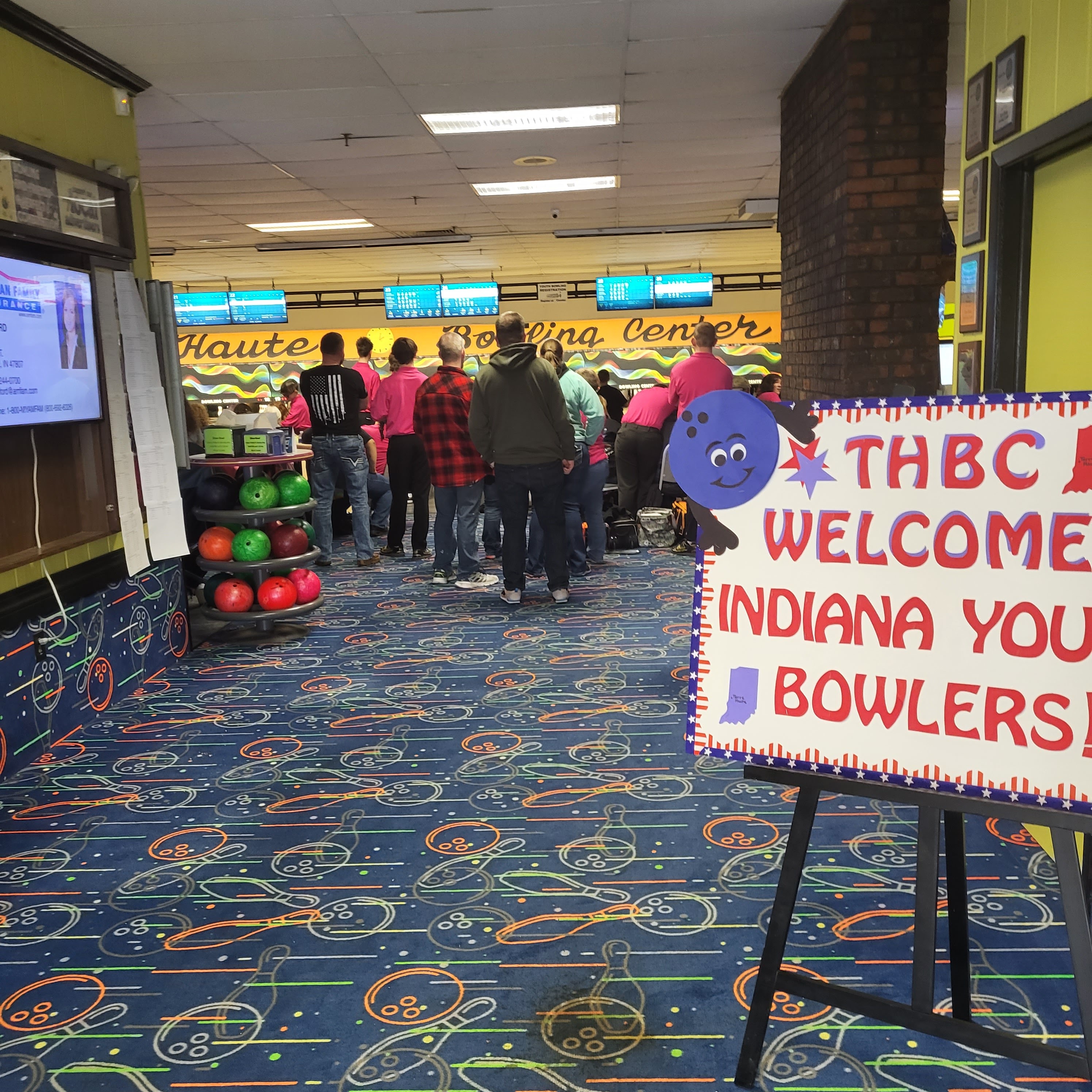 A welcome sign for Indiana youth bowlers stands in a bowling alley concourse with people and lanes in the background.