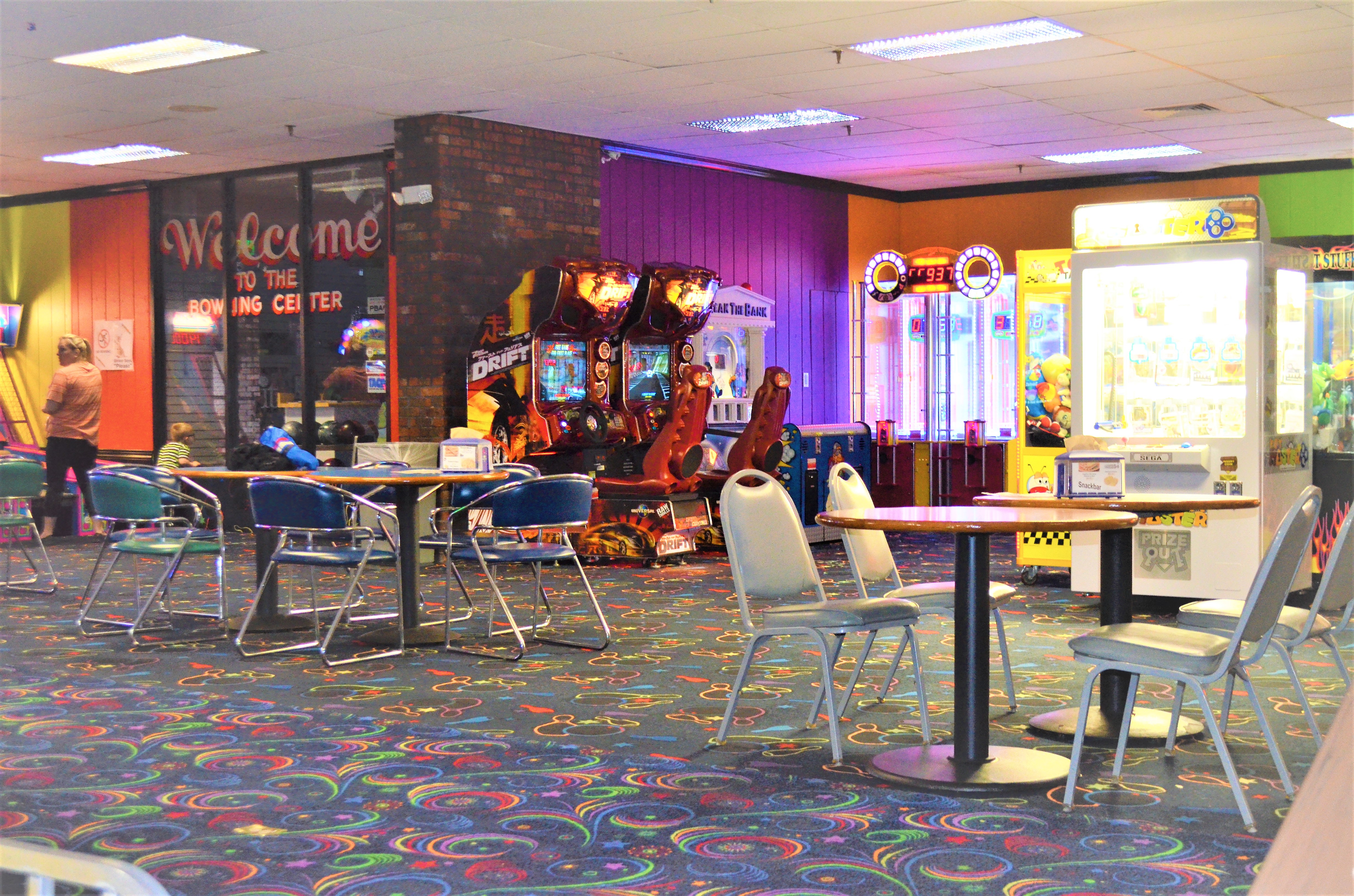 An arcade area inside a bowling center featuring driving games, claw machines, and several round tables with chairs.