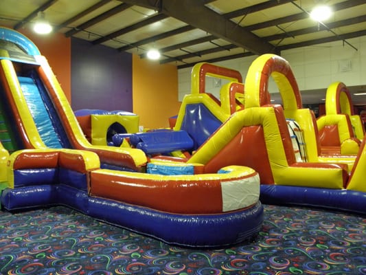 Large colorful inflatable slides and obstacle courses in an indoor playground with patterned carpet.