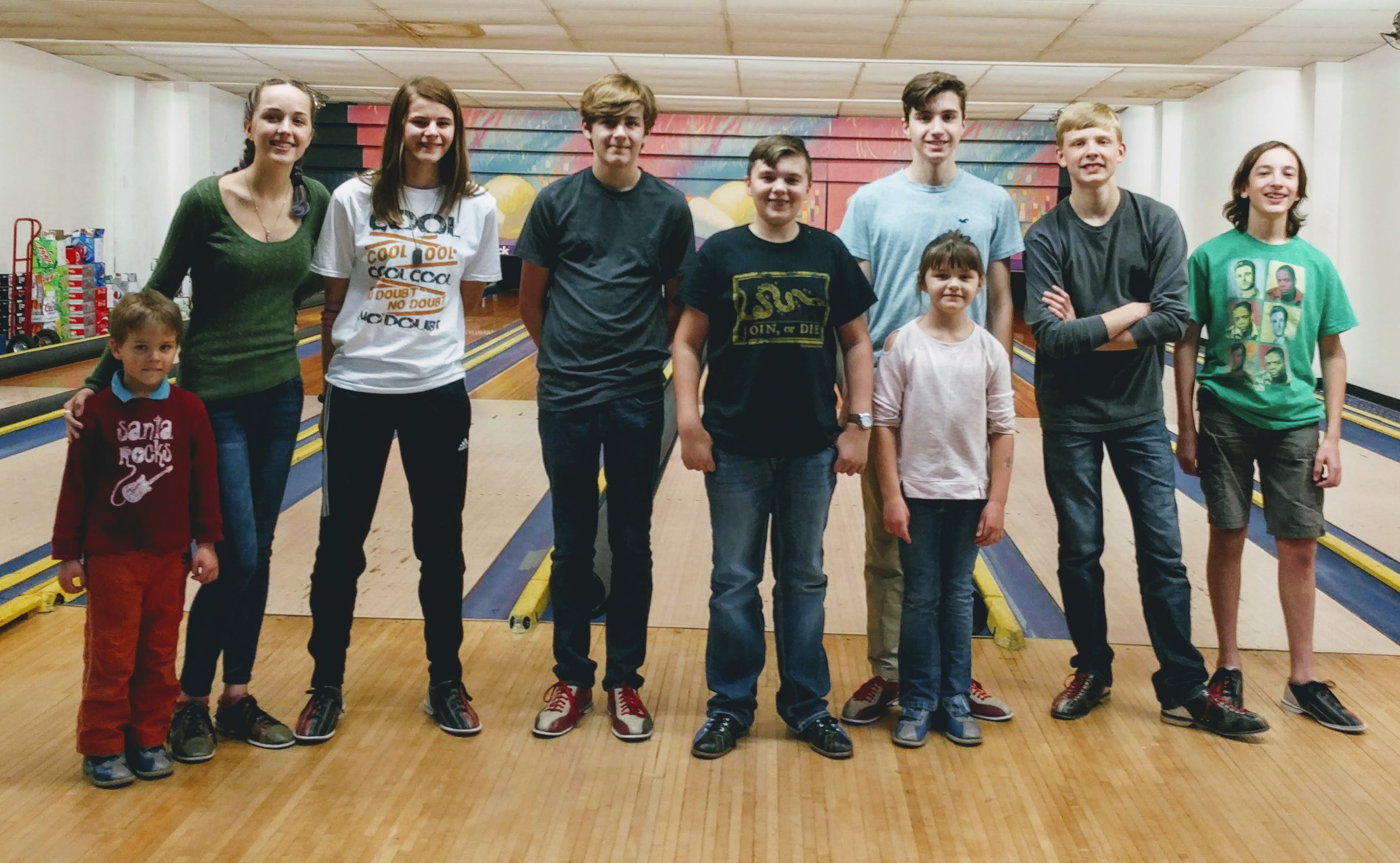A group of nine smiling children and young adults stand side-by-side on a bowling lane approach area.