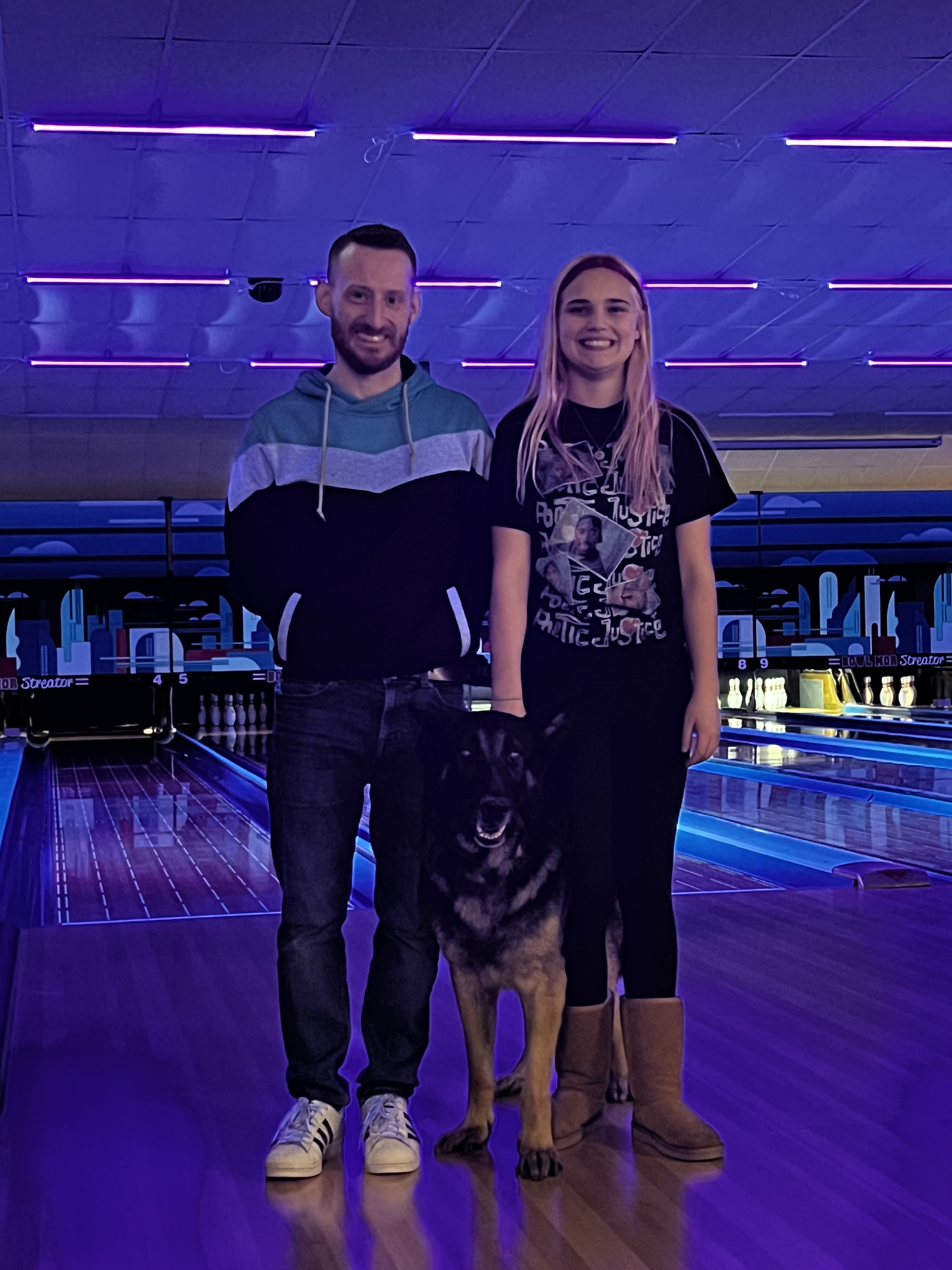 A man, a woman, and a German Shepherd stand on a bowling lane approach under purple and blue cosmic lighting.