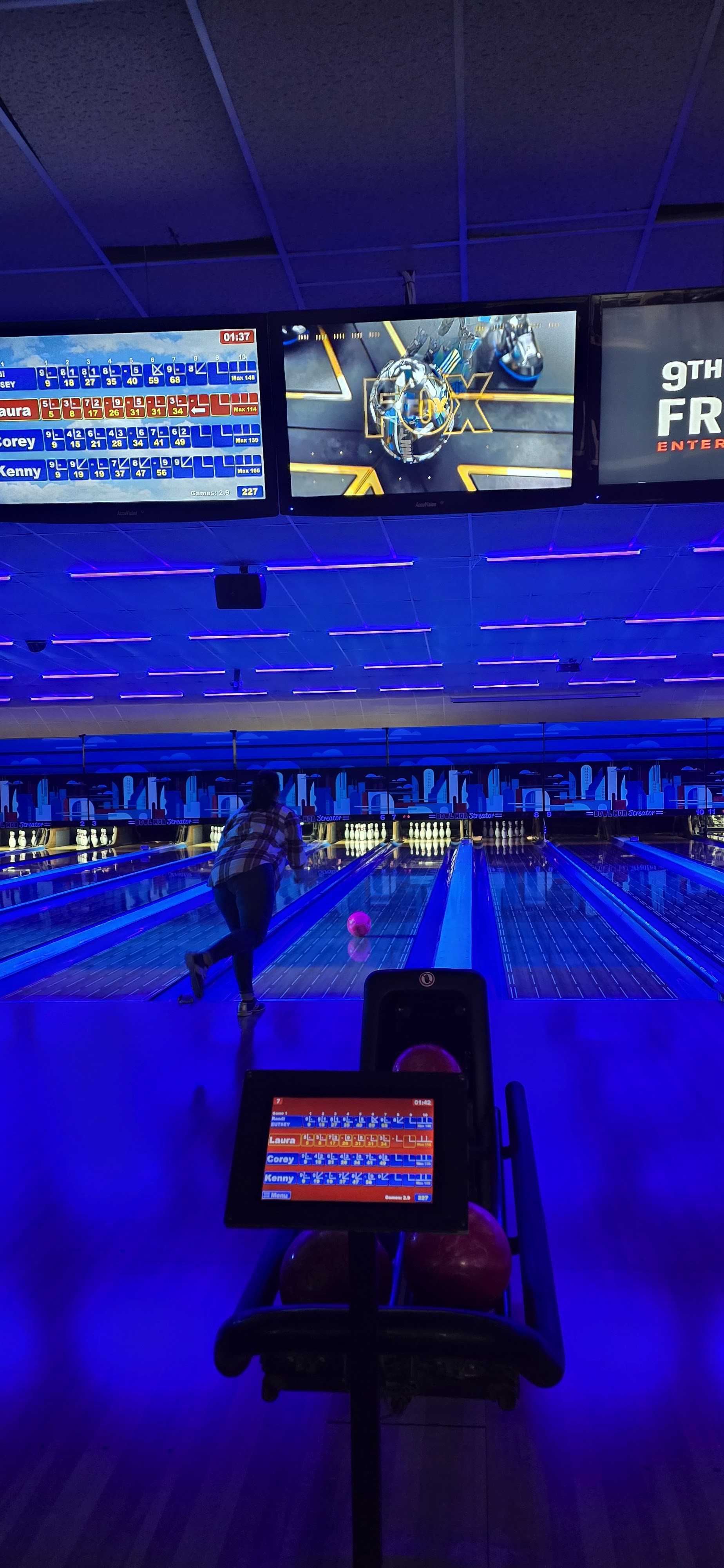 A person bowls on a lane under blue cosmic lighting with digital scoreboards visible overhead.