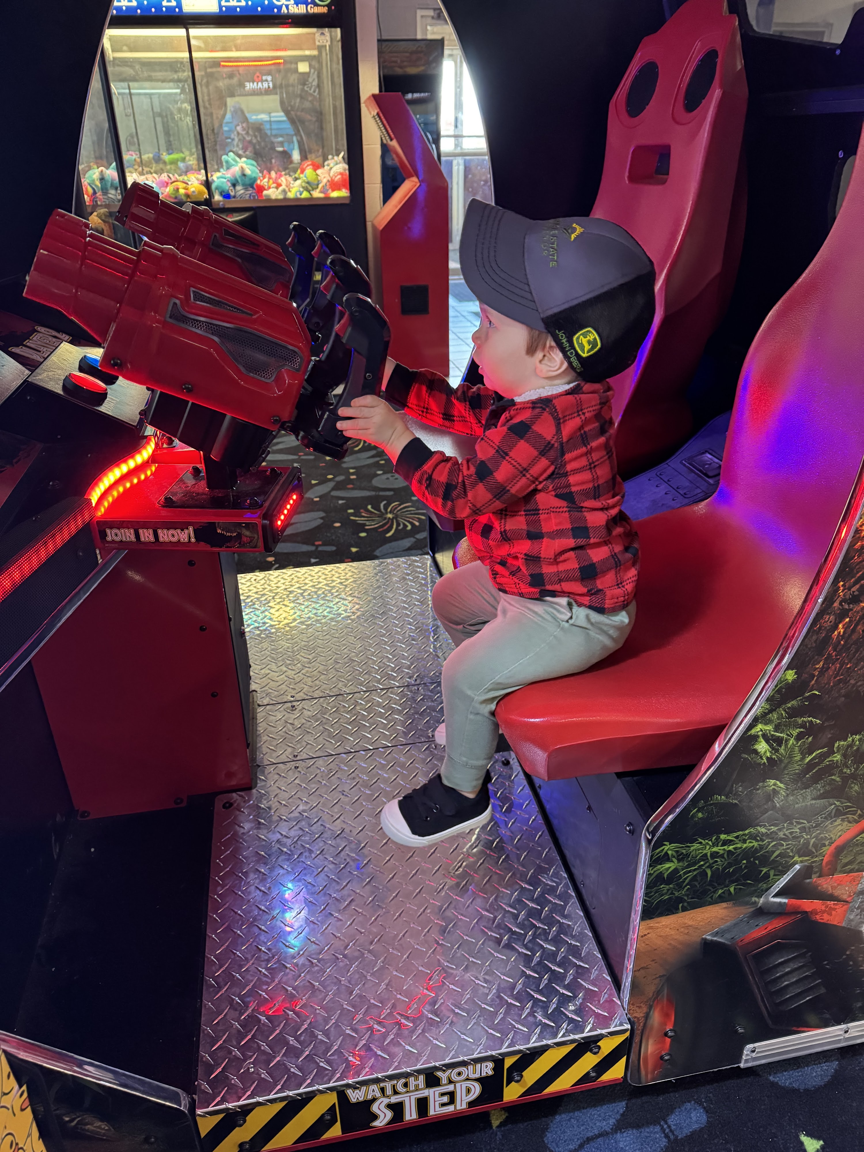 A young child in a plaid shirt and cap plays a red sit-down arcade game in a colorful game room.