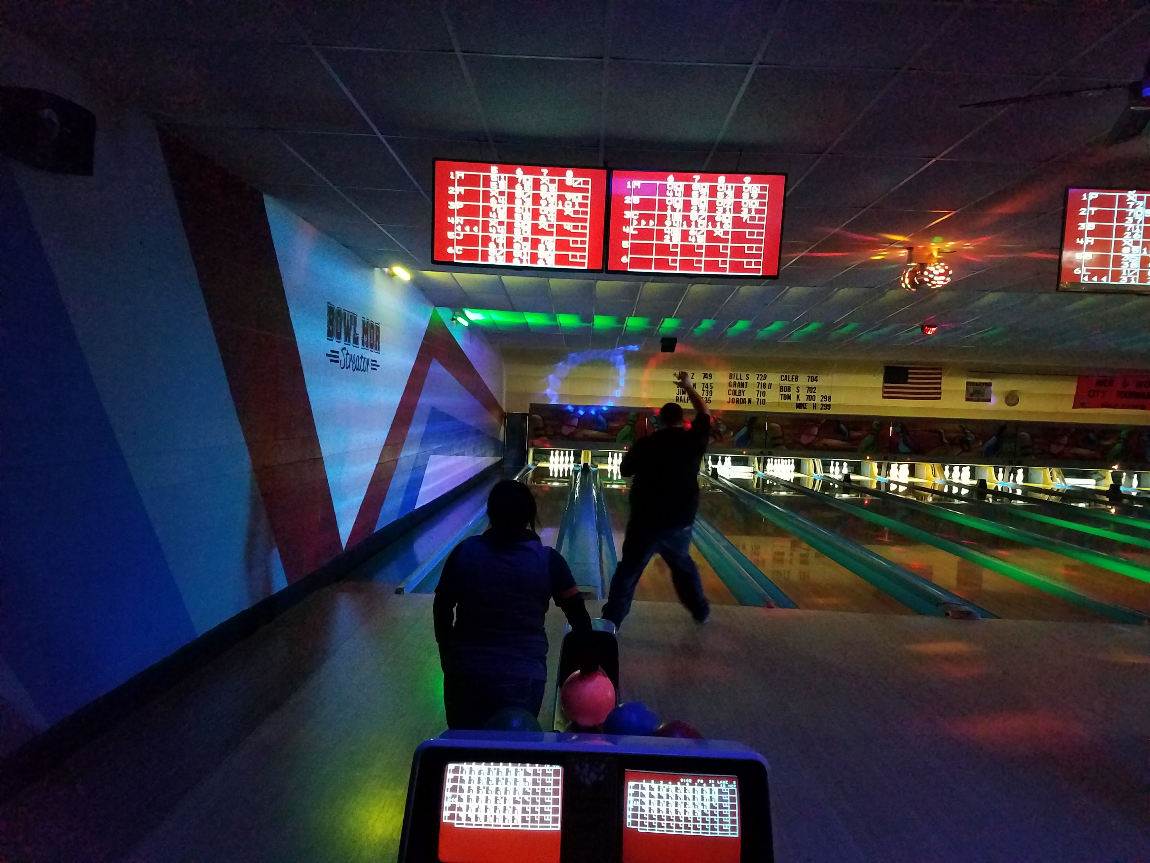 Two people bowling at night in an alley with neon blue, green, and red cosmic lighting and electronic scoreboards.