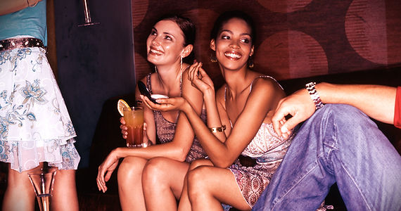 Two women smiling and holding cocktails while sitting on a couch in a social lounge setting.