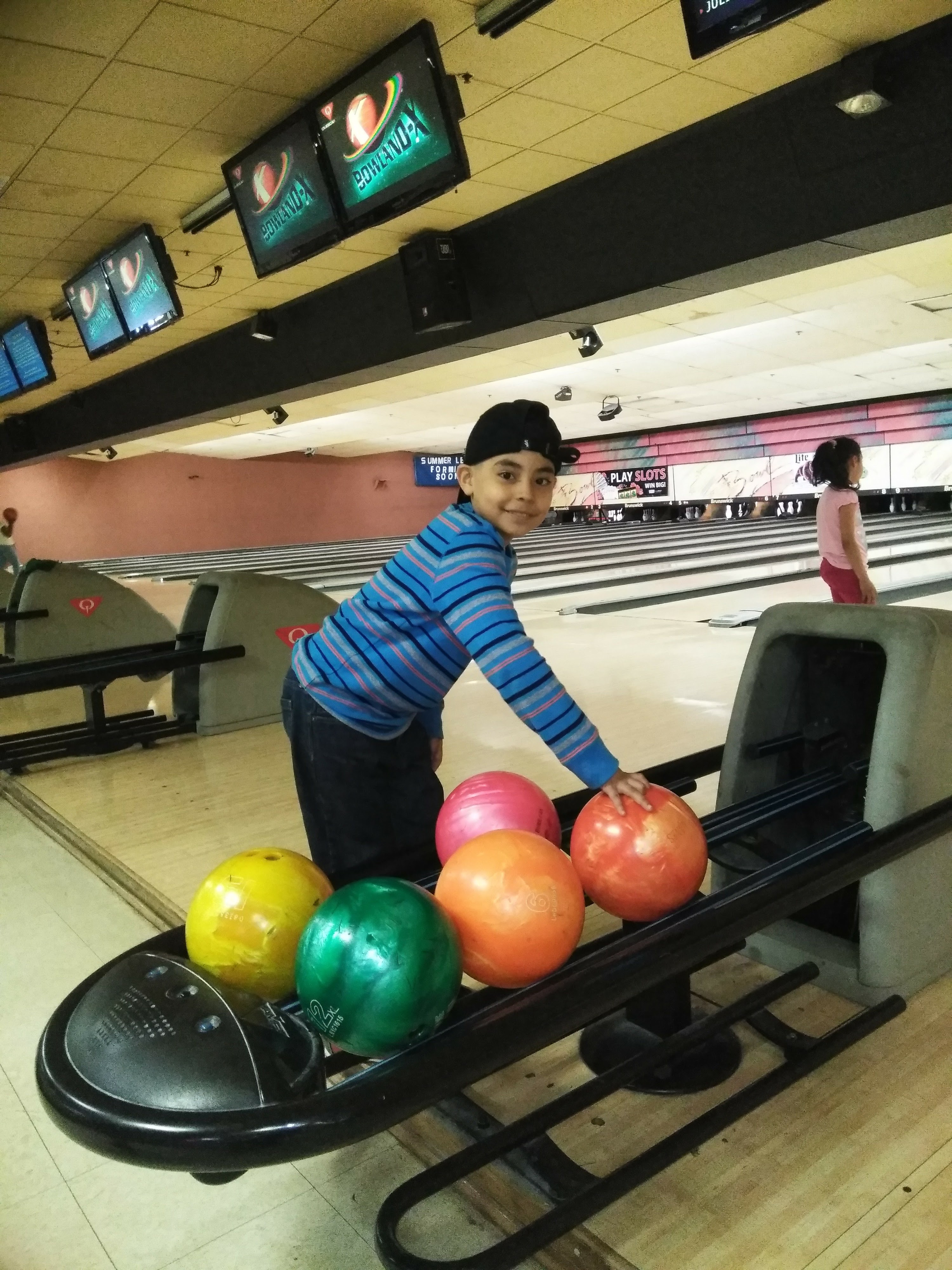 A smiling young boy in a blue striped shirt and black hat poses by colorful bowling balls at a bowling alley.