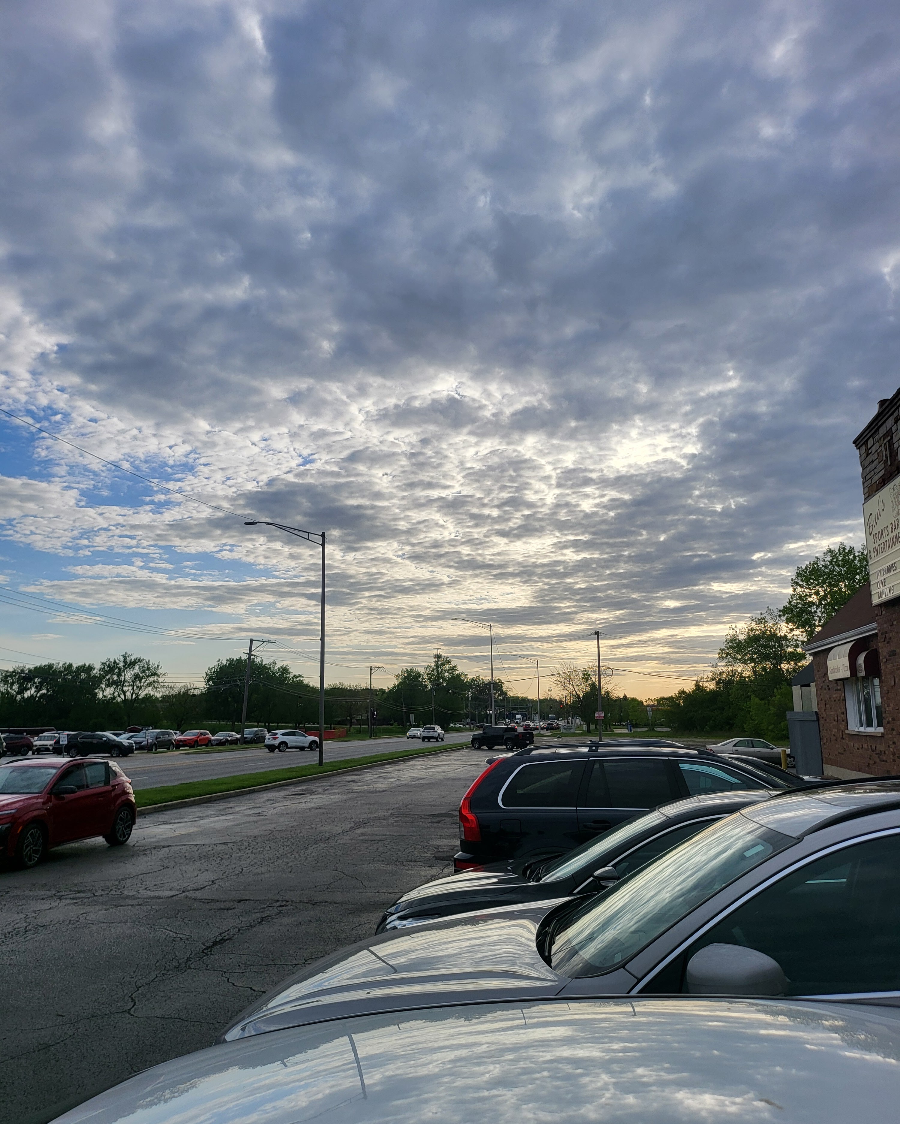 An outdoor parking lot with cars under a wide, cloudy sunset sky next to a brick building.