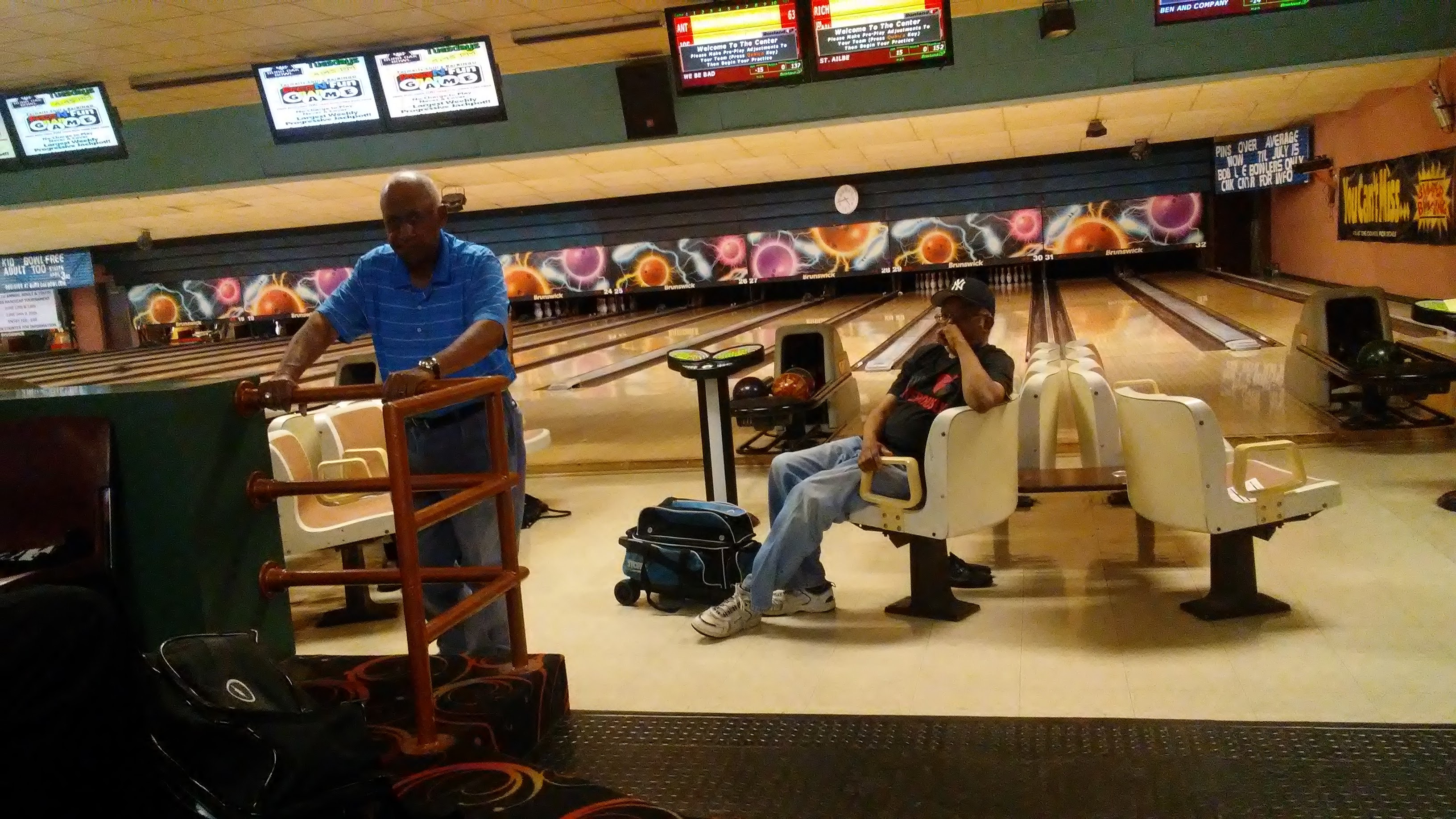 Two men at a bowling alley with wooden lanes and overhead scoring monitors in a well-lit interior.
