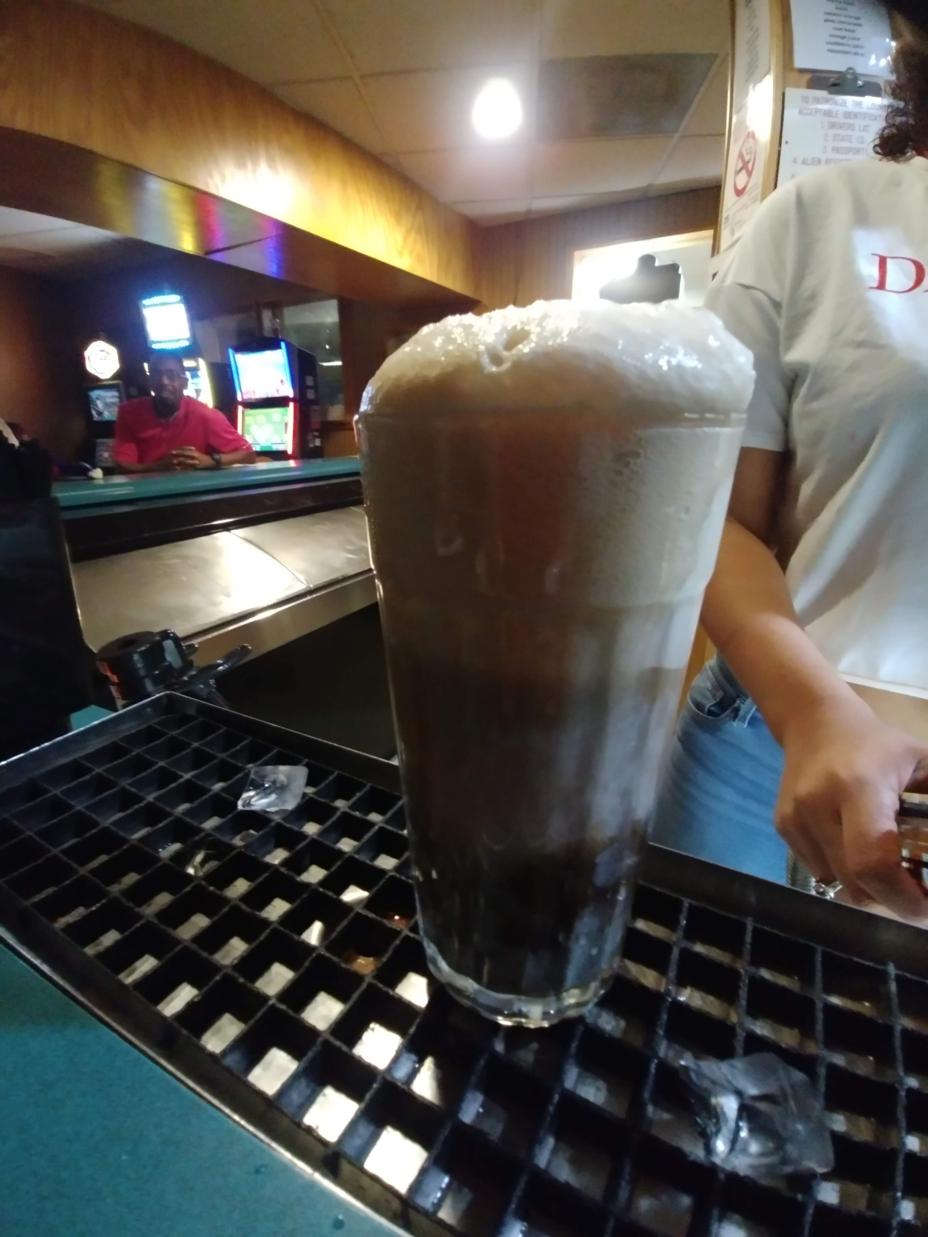 A frothy root beer float in a tall glass sits on a black bar mat inside a bowling alley lounge.
