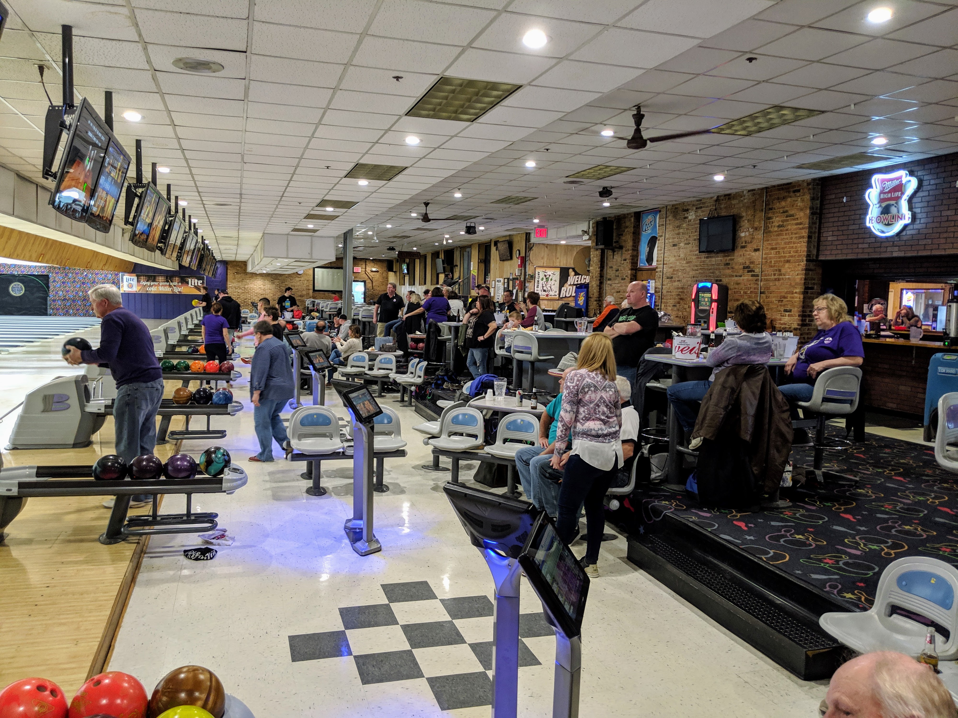 Interior view of a lively bowling center with people playing on several lanes and a bar area in the background.