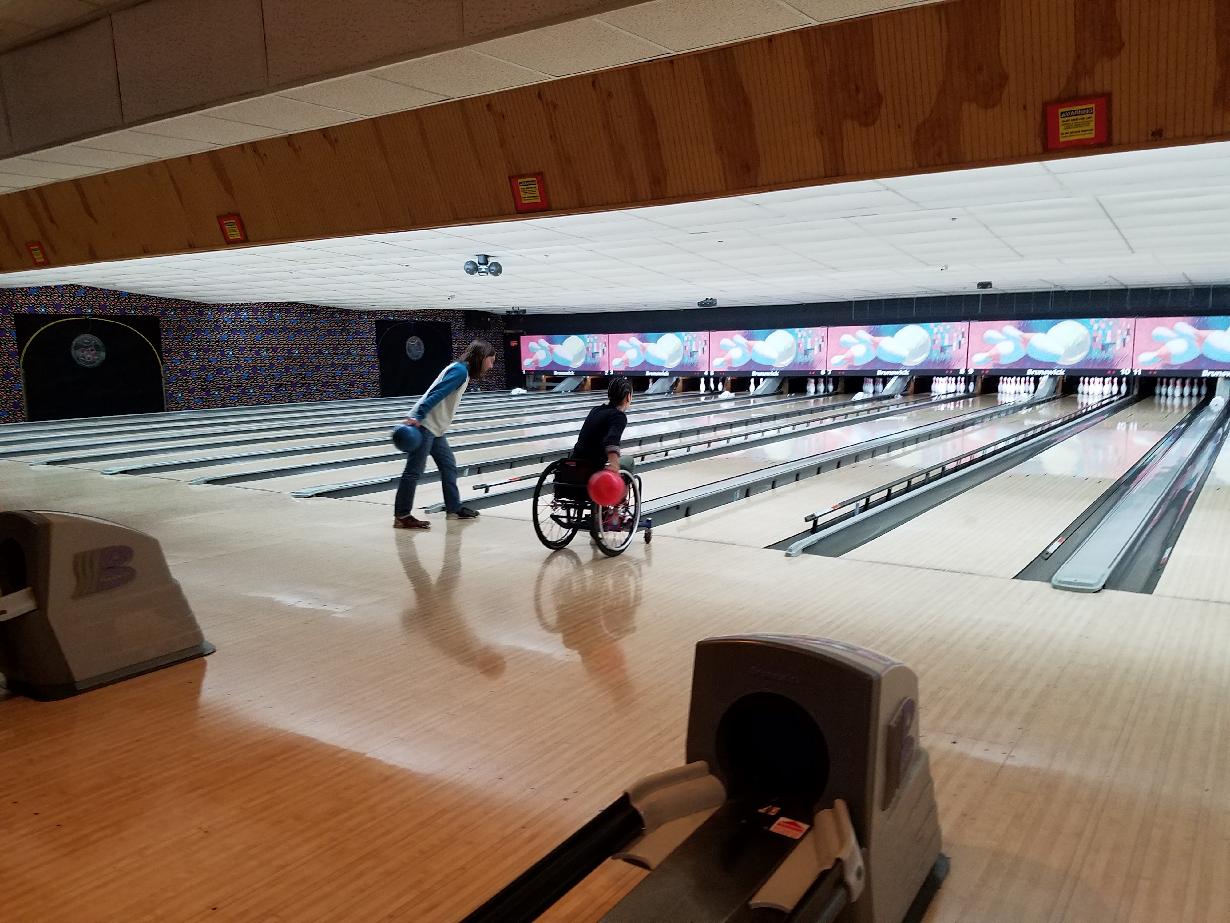 A person in a wheelchair and a standing person bowling side-by-side at a well-lit bowling alley.