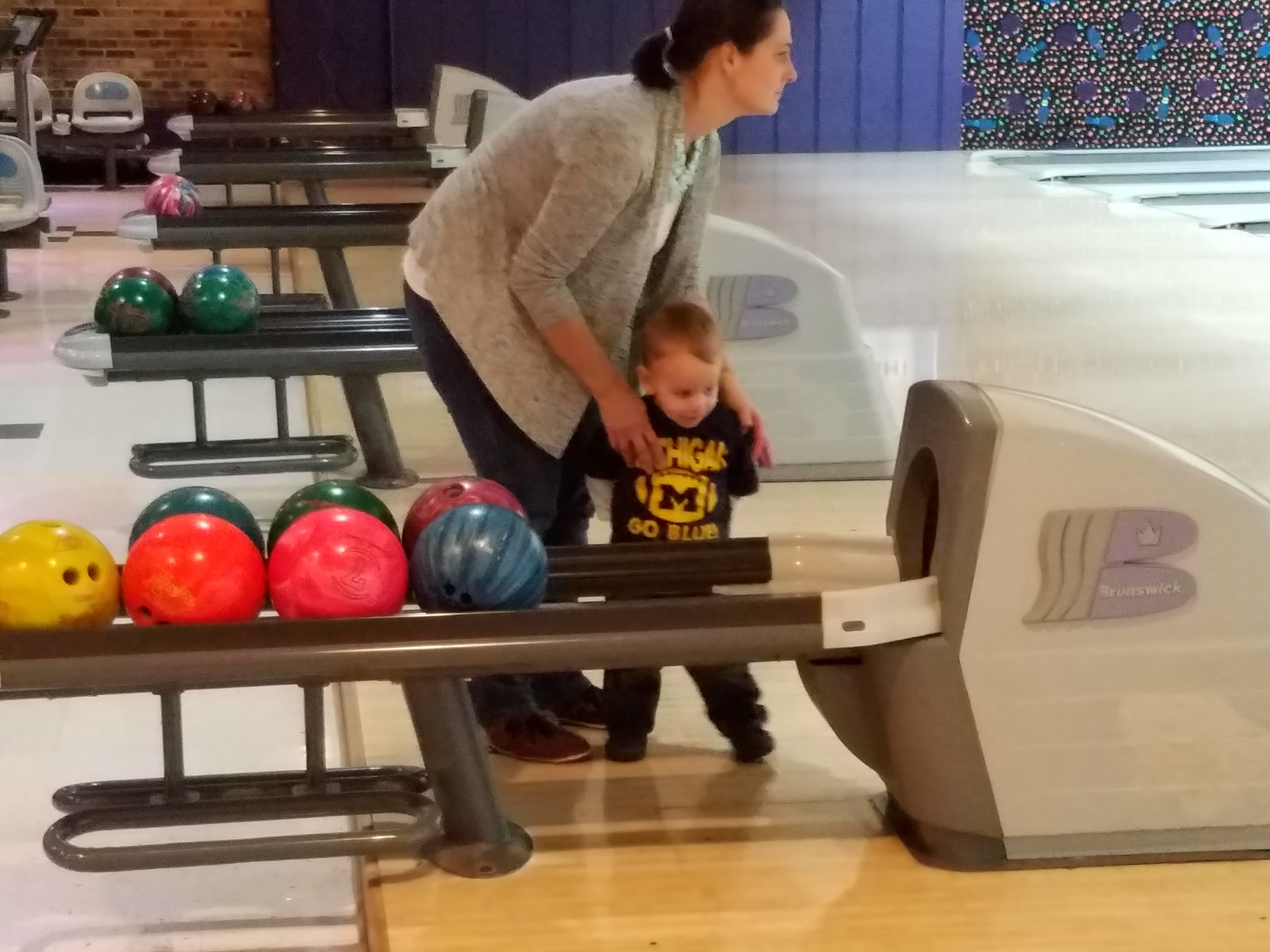 A woman assists a young child standing next to a bowling ball return rack at a bowling center.