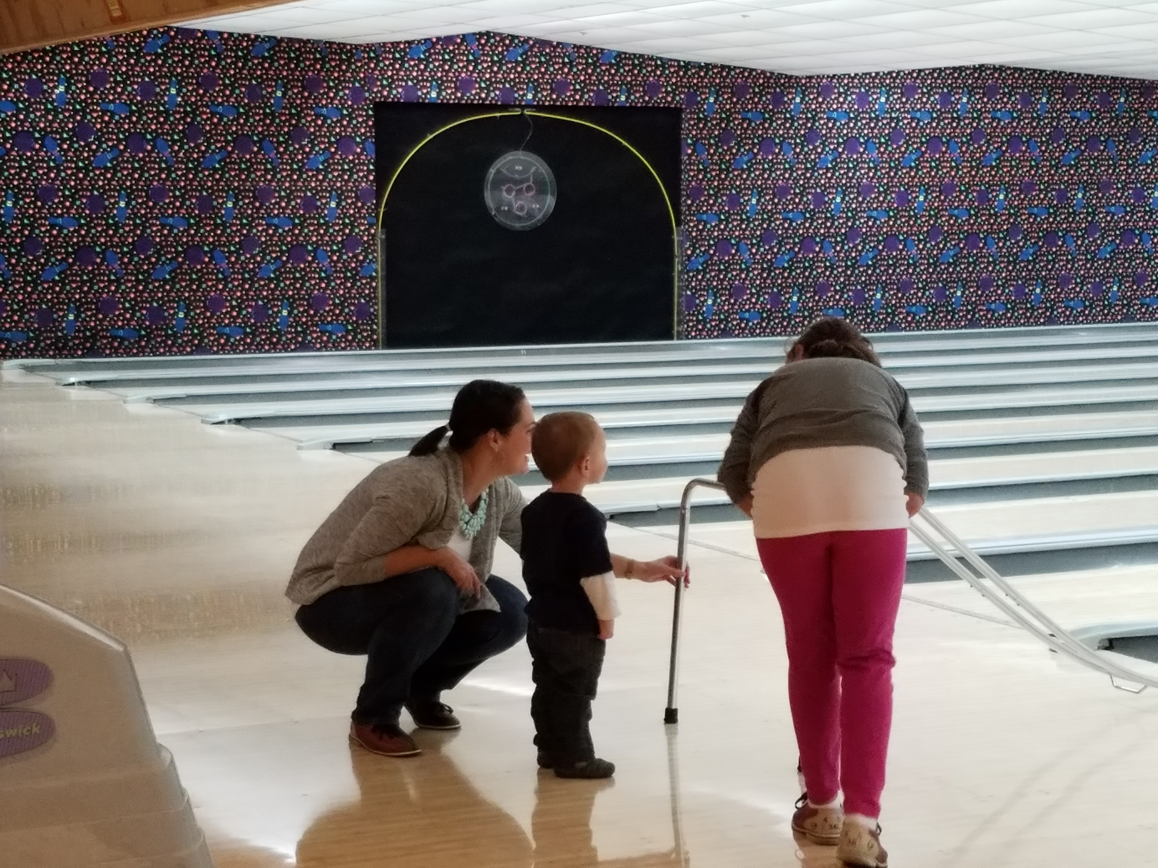 A woman and a young child watch as another person uses a bowling ramp on the lanes.