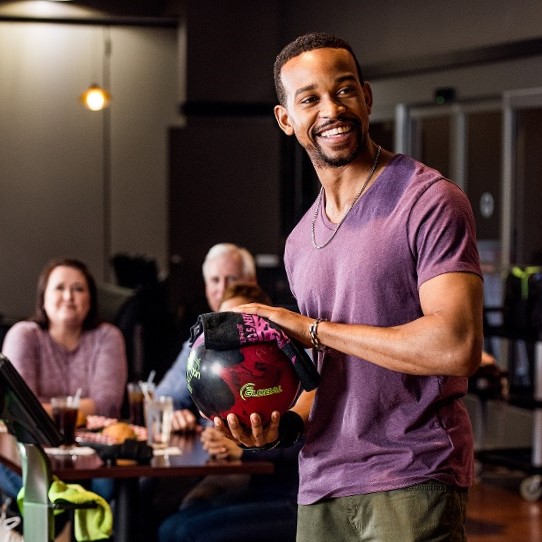 A man in a purple shirt smiles while holding a red and black bowling ball at a bowling alley.