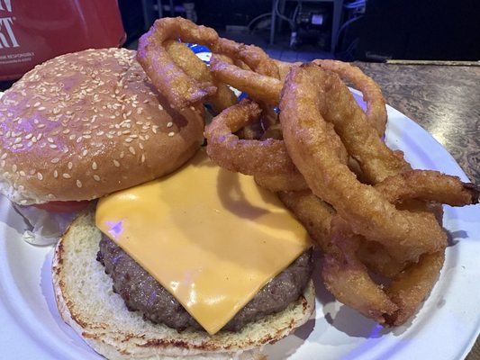 A cheeseburger with a sesame seed bun and a side of crispy fried onion rings on a white plate.