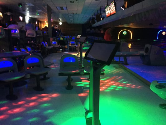 A dimly lit bowling alley with neon blue chair backs and colorful light patterns projected on the floor.