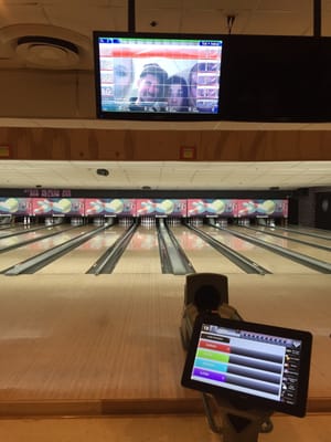 A view of a bowling lane featuring an interactive scoring tablet and an overhead monitor showing player photos.