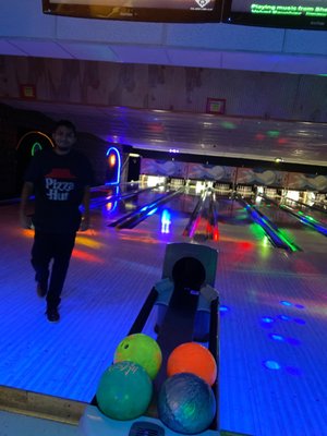 A person stands on a bowling lane approach under blue cosmic lighting with colorful bowling balls in the foreground.