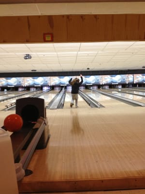 A child in a dark jacket celebrates with arms raised on a bowling lane with bumpers.