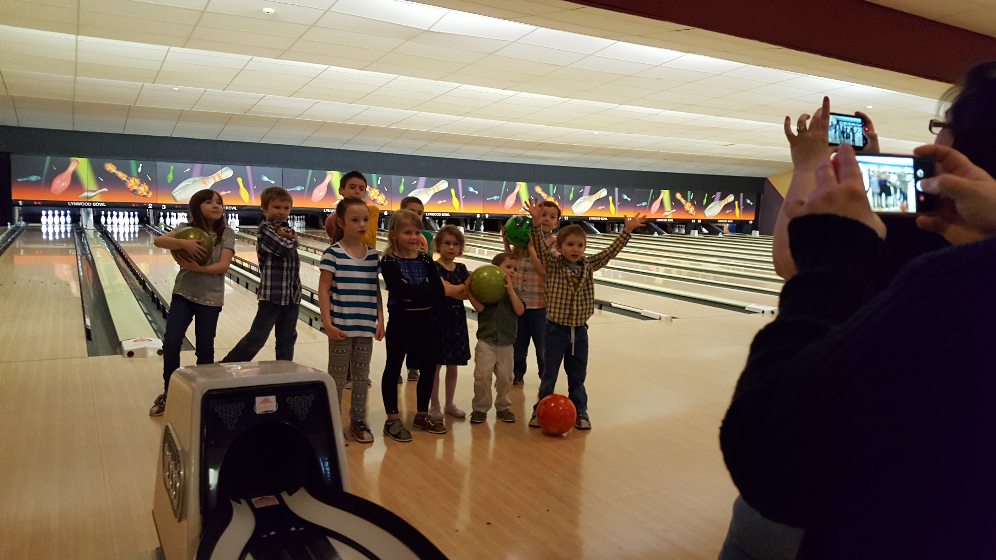 A group of happy children posing with bowling balls on a lane while an adult takes their picture.