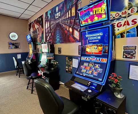 A row of illuminated video gaming terminals with black chairs in a lounge area.