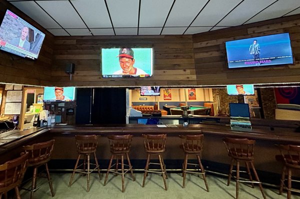 An empty bar with wooden stools and multiple televisions mounted on wood-paneled walls.