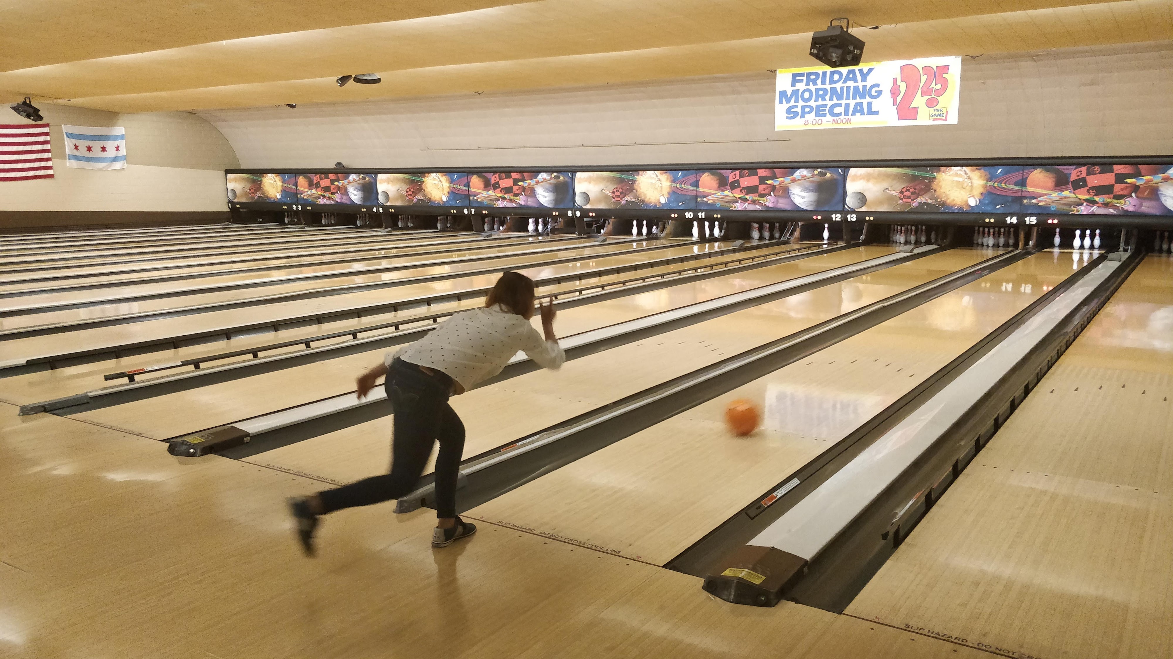 A woman bowls an orange ball down a lane at a well-lit Chicago bowling alley with flags on the wall.